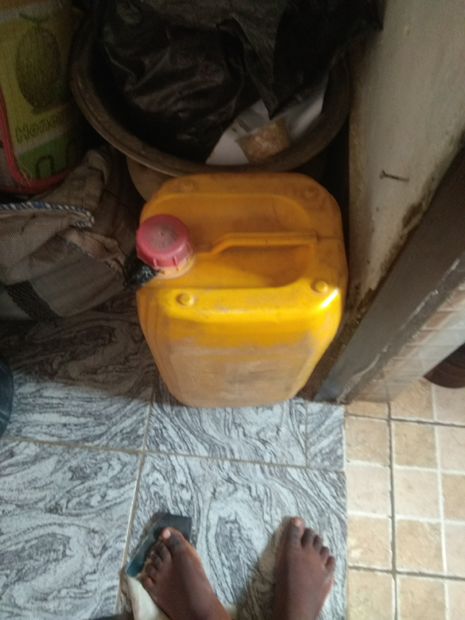 The image shows a close-up view of a yellow plastic container with a red cap, placed on the floor. The container appears to be a jerrycan, possibly holding liquid. The floor is tiled with a pattern of grey and white, transitioning to beige tiles. In the foreground, a person's bare feet are visible, with one foot positioned next to a dark object that might be a rag or a cleaning cloth. The feet suggest the person is standing or kneeling.
Behind the jerrycan, a large black plastic bag is visible, contained within a round, dark basin or bowl. To the left of the jerrycan, there is a partially visible yellow bag with the word "Honey" printed on it in black letters, along with an illustration of a fruit, possibly a melon. Some textured fabric or material is also seen to the left, draped over something.
The setting appears to be indoors, possibly a storage area or a living space with utilitarian items. The lighting suggests it might be daytime, though no windows or direct light sources are clearly visible. The overall impression is of a simple, possibly domestic, environment in Jalingo, Nigeria. No specific activity or event is clearly depicted, other than the implied presence of the person whose feet are visible. The image is focused on the arrangement of objects and the immediate surroundings.