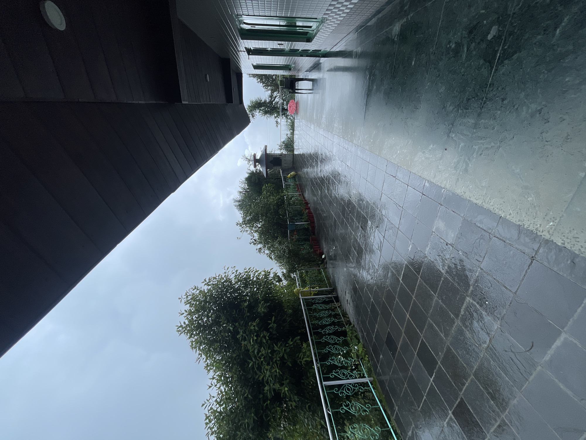The image shows a wet, tiled balcony or veranda of a building, likely a hotel or guesthouse, in Bhagsu, India. The sky is overcast and a light rain appears to be falling, evident from the reflections and water on the dark gray, square tiles of the balcony floor. To the left, a metal railing with a decorative pattern runs along the edge, beyond which lush green trees and foliage are visible, suggesting a mountainous or hilly terrain. In the background, a small, stone structure with a tiered roof stands amidst the greenery, and further out, the misty outlines of hills or mountains can be seen. The right side of the image shows the exterior wall of the building, clad in white and grey square tiles, with green-framed windows. A black chair and a pink circular object, possibly a cushion or mat, are placed on the balcony. The overall atmosphere is serene and a bit melancholic due to the weather.