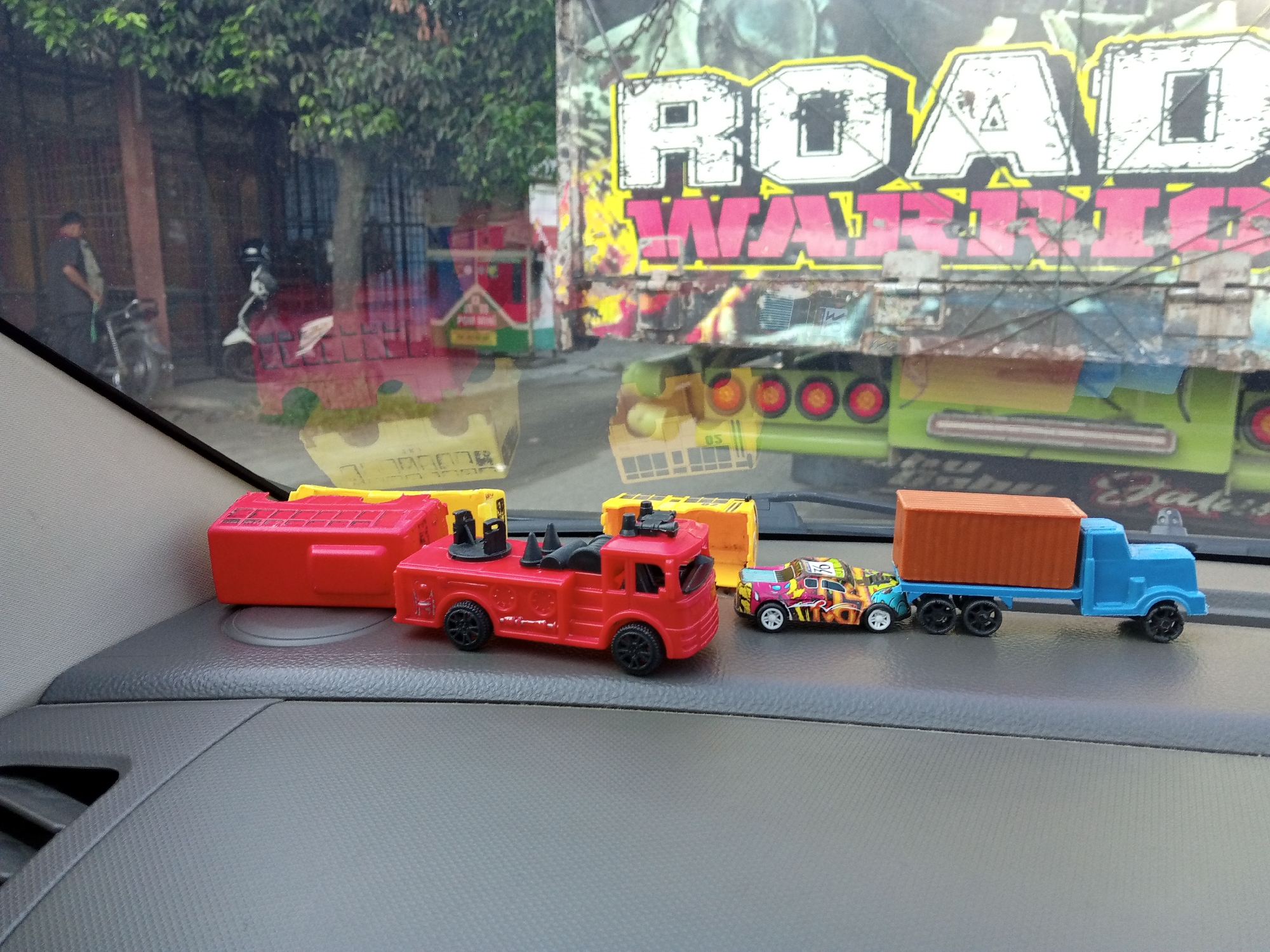 In Lhoknga, Indonesia, a toy fire truck sits on the dashboard of a vehicle. Behind the toy, a yellow box is visible, and further back, a larger, red toy truck is present. The scene is viewed through a vehicle's windshield, reflecting the exterior. Outside, a large truck with the words "ROAD WARRIOR" emblazoned on its side is parked, along with other vehicles and buildings. A person in a dark shirt and a helmet is visible near a motorcycle. Trees and foliage are also present in the background.