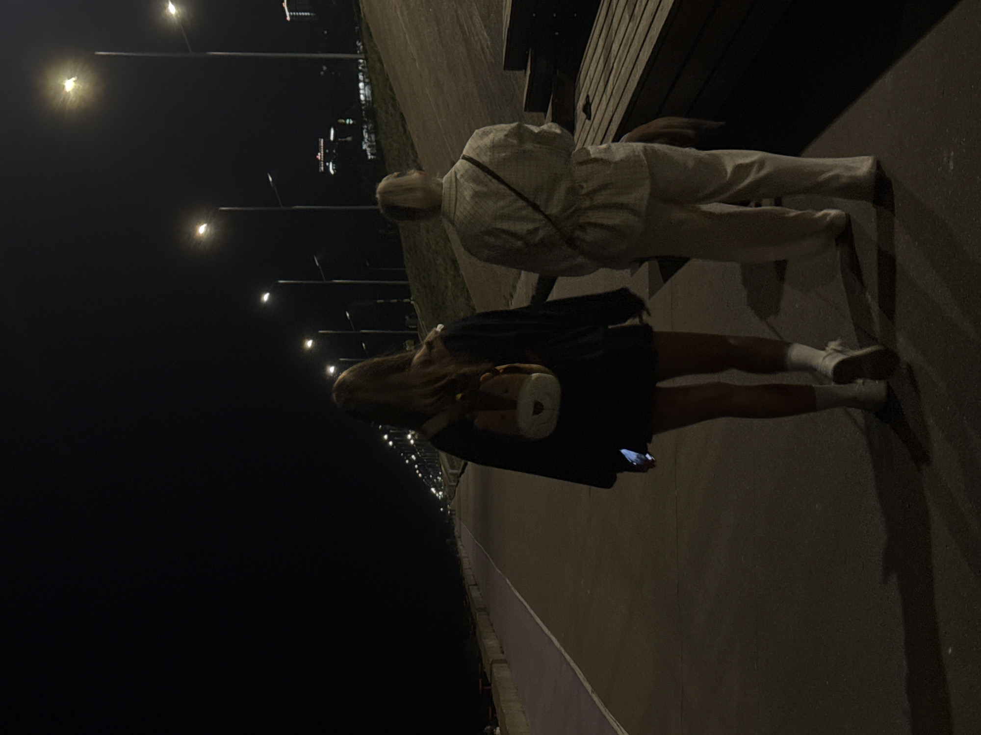 A nighttime photo shows two people on a paved walkway. One person sits on a bench, wearing light-colored pants and a patterned top. The second person walks, wearing a dark dress, socks, and sneakers, holding a cylindrical object in one hand and a phone in the other. Street lamps illuminate the scene, with taller buildings visible in the distance. This is in Nizhny Novgorod, Russia.