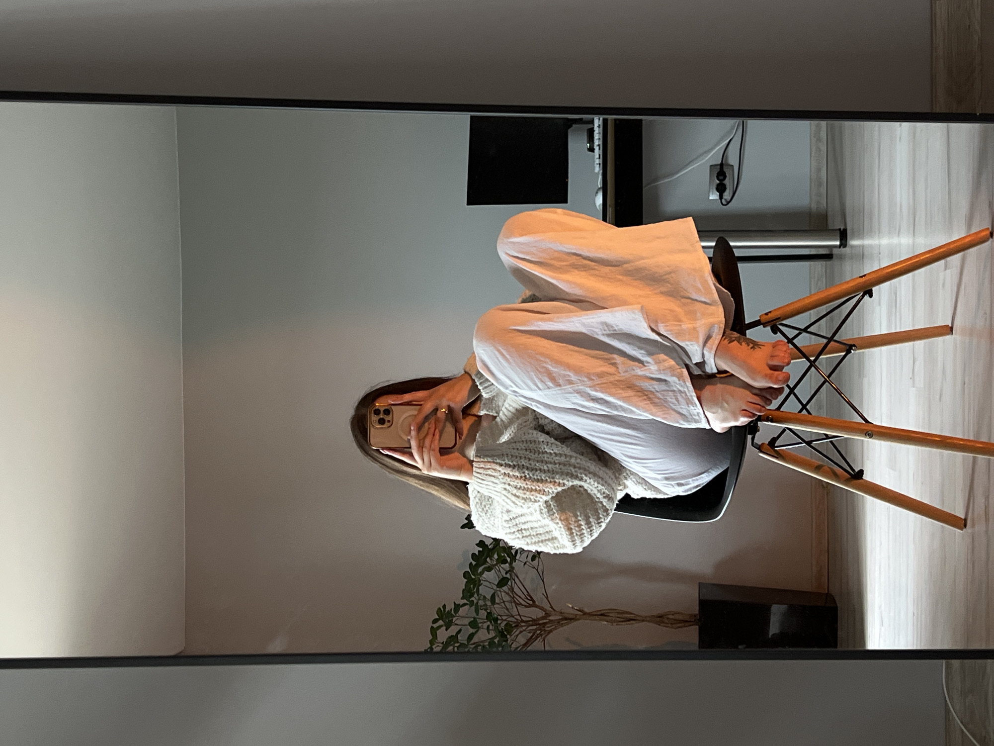 A mirror selfie captures a young woman seated casually in a modern, minimalist room. The image is a reflection in a large, dark-framed mirror, showing the woman from approximately the waist up, though her legs are visible. She has long, straight, light brown hair and holds a light-colored smartphone, obscuring her face, as she takes the photo. She is wearing an oversized, chunky knit, cream or white sweater and light-colored, wide-leg pants. Her bare feet, one with a small dark tattoo on the top, are resting on the seat of the chair. A simple ring is visible on her right hand.
The chair is a contemporary design with a black molded seat and natural wood legs supported by black wire. Behind the woman, inside the reflection, is a light-colored wall. To her left (viewer's right in the mirror), a small leafy indoor plant with intricate branches is placed on a surface. Further back, a dark desk holds a black computer monitor, and electrical cables are visible on the wall, leading to an outlet. The room features light-colored walls and light-toned, wood-look flooring. The lighting appears soft and natural, suggesting daytime. No discernible text is visible. The scene depicts a relaxed moment in a stylish indoor setting, located in Ternopil, Ukraine, though no specific location cues are visible in the image itself.