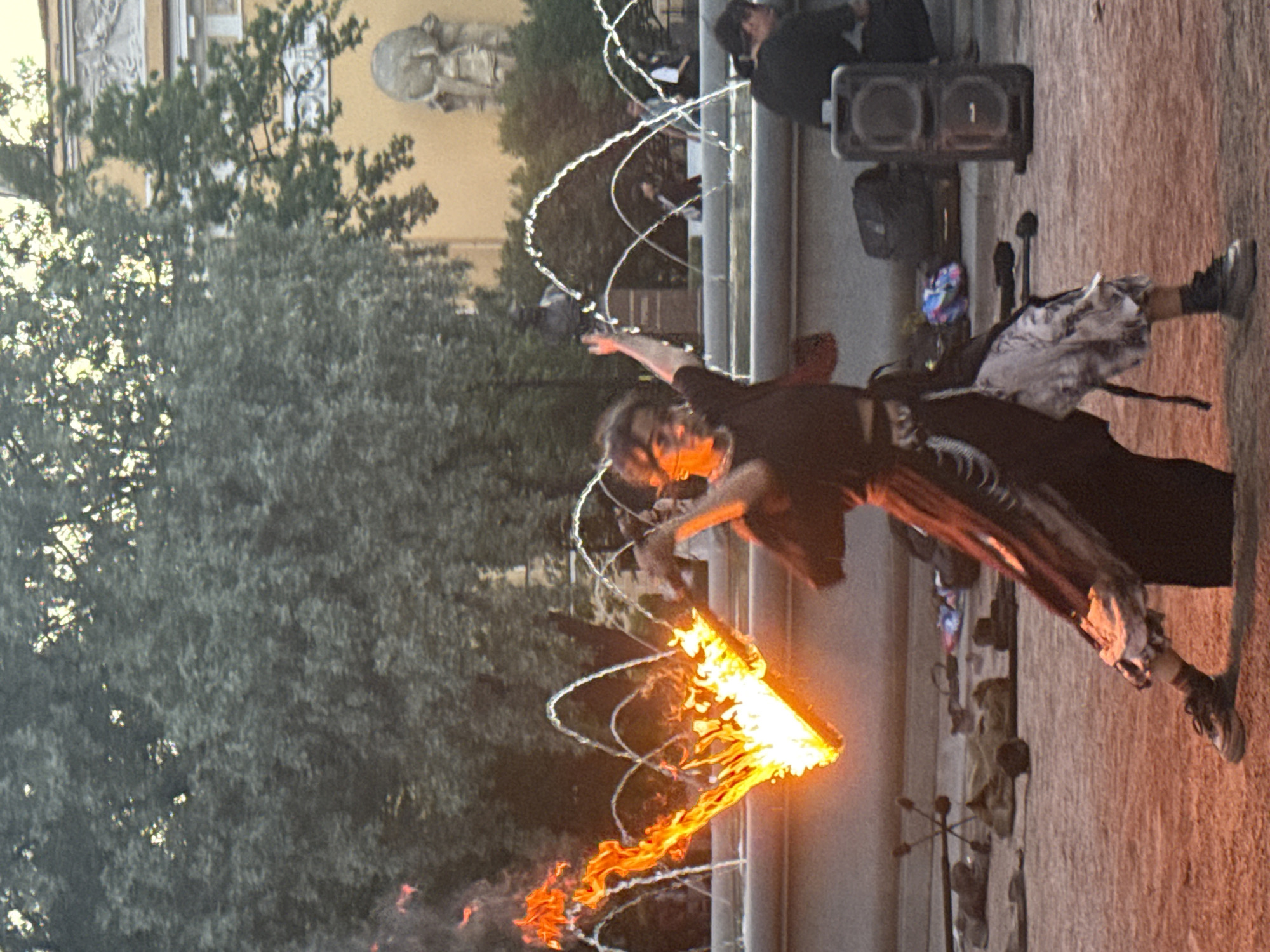 A person is performing a fire dance in front of a fountain in Saint Petersburg, Russia. The person, dressed in dark clothing and a long skirt, is spinning a prop that is engulfed in flames, creating a dynamic display of fire. The fountain is spraying water into the air, with arched streams creating a backdrop to the performance. In the background, trees and a building with classical architectural details are visible. Several other people are present, some observing the performance and others seated near a speaker system. The ground is a dusty, sandy surface. The overall scene depicts an outdoor urban entertainment event, likely in a public square or park.