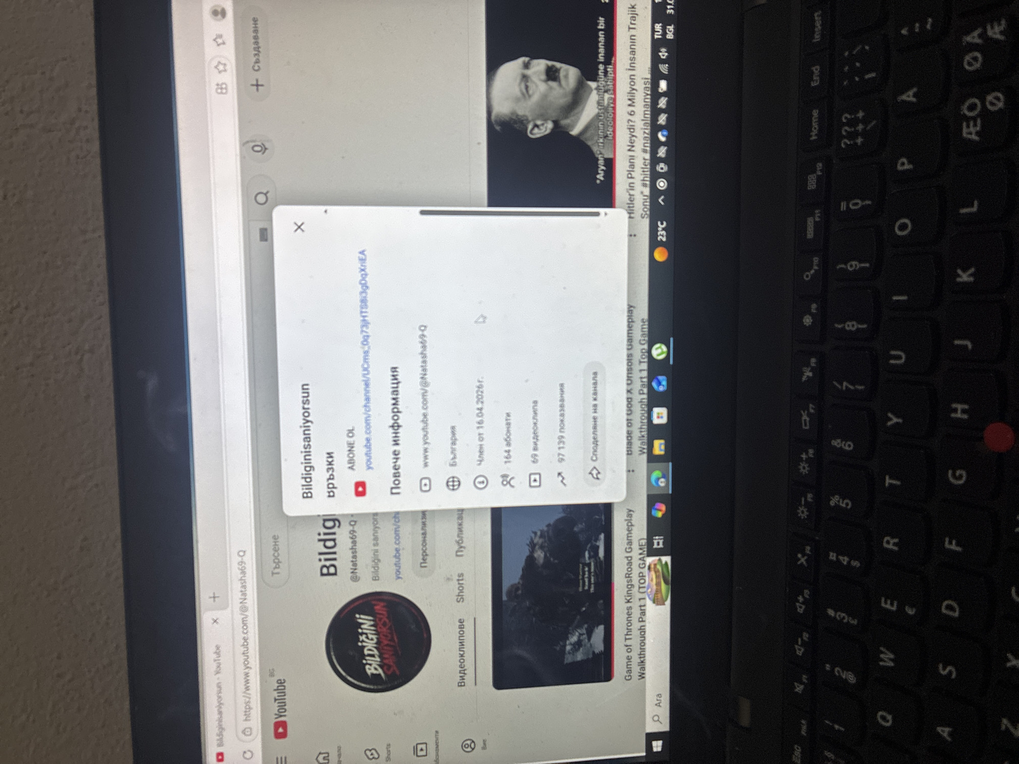 The image shows a laptop screen displaying a YouTube channel page named "Bildiginisaniyorsun" (translation from Turkish: "Do you think you know?"). The channel's profile picture is visible along with statistics like 164 subscribers and 69 videos. A popup window is partially obscuring the YouTube page, showing options to view the channel URL and other information. Above the YouTube page, a tab displays a video featuring Adolf Hitler with a Turkish title translating to "What was Hitler's plan? 6 Million People's Tragic End." Below the YouTube page, the taskbar shows various programs, including a game called "Game of Thrones KingsRoad Gameplay." The laptop's keyboard is visible in the foreground. The location is not identifiable from the image.