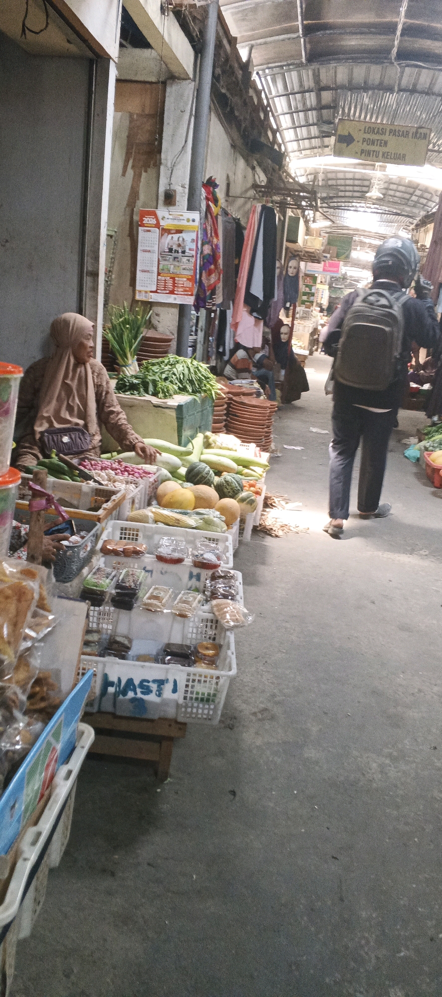The image captures an outdoor market scene in Maduran, Indonesia. A vendor wearing a hijab sits behind a stall overflowing with fresh produce, including gourds, cucumbers, radishes, and leafy greens. Baskets of packaged goods, possibly snacks or condiments, are neatly arranged on a lower shelf. A man wearing a helmet and a backpack walks away from the camera down a paved walkway lined with market stalls. Overhead, a corrugated metal roof provides shade. A directional sign points towards "LOKASI PASAR IKAN" (Fish Market Location), "PONTEN" (likely a location name), and "PINTU KELUAR" (Exit Door). The overall impression is that of a bustling local marketplace.