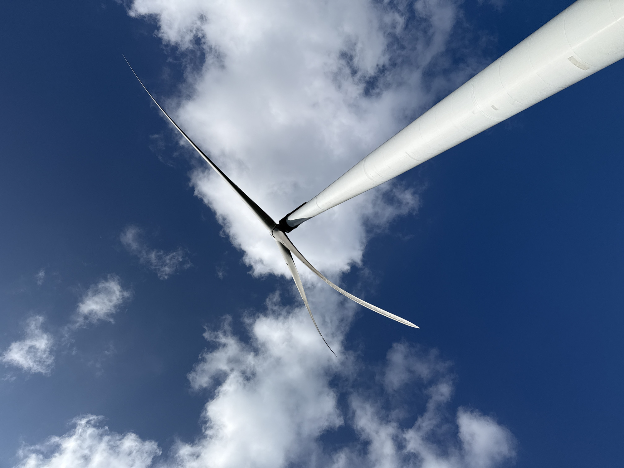 An extreme low-angle shot captures a white wind turbine against a vibrant blue sky dotted with fluffy white clouds. The turbine's tall, cylindrical tower extends upwards diagonally from the lower right corner of the frame. At the top of the tower, three blades are visible, angled upwards and to the left. The blades appear sleek and modern, with one prominent in the foreground, and the other two slightly further back and angled differently. The clouds are scattered, with some larger formations on the left and smaller wisps dispersed throughout the sky. The lighting suggests a bright, sunny day. The image does not depict any people or specific architectural details that would indicate a precise urban location within Trường Thọ, Vietnam.