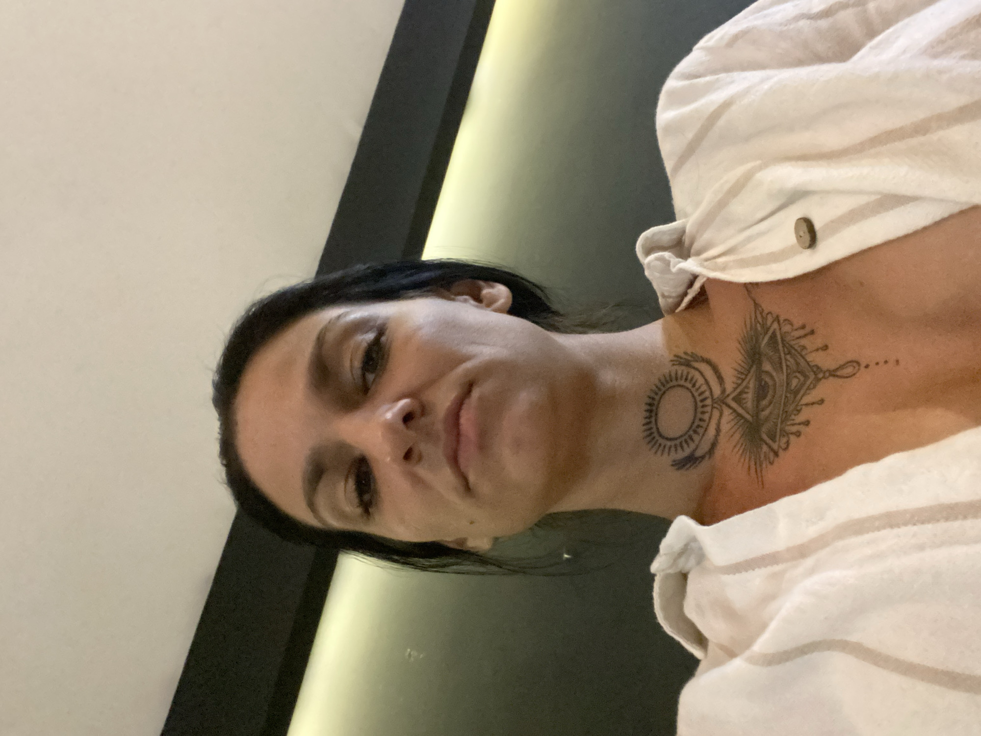 A selfie-style photograph presents a woman with dark hair and light skin from a low angle. The background consists of a light-colored wall adjacent to a black frame or molding, with a subtle yellow light strip between the wall and frame. The woman is wearing a striped, light-colored shirt. Prominent tattoos are visible on her neck and upper chest: a circle with rays resembling a sun and an eye within a geometric shape. No other discernible features indicate a specific location or activity.