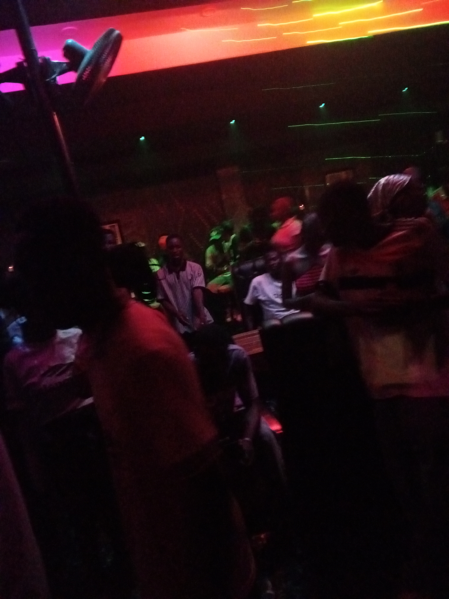 Location: Unknown, Unknown, a dimly lit indoor setting with multiple people.
People: Several individuals are present, some standing, some sitting. A person wearing a red shirt is prominently visible in the foreground. In the background, at least two people are sitting down, one in a white shirt, and another in a striped shirt. Some individuals are blurred or partially obscured by darkness.
Subjects: The subjects include people and possibly furniture or other objects within the room. The lighting appears to be artificial, with colorful lights illuminating the scene.
Objects: A fan-like object on a stand is visible in the left foreground. There are also seating arrangements, likely chairs, visible in the background.
Actions: People are present and appear to be socializing or relaxing.
Interactions: The image suggests interactions between individuals, although the low light and blurriness make it difficult to determine specifics.
Environment: The environment is dark, with artificial lighting creating a nightclub or lounge-like atmosphere. The overall impression is that it is a social gathering in an indoor space with limited lighting.