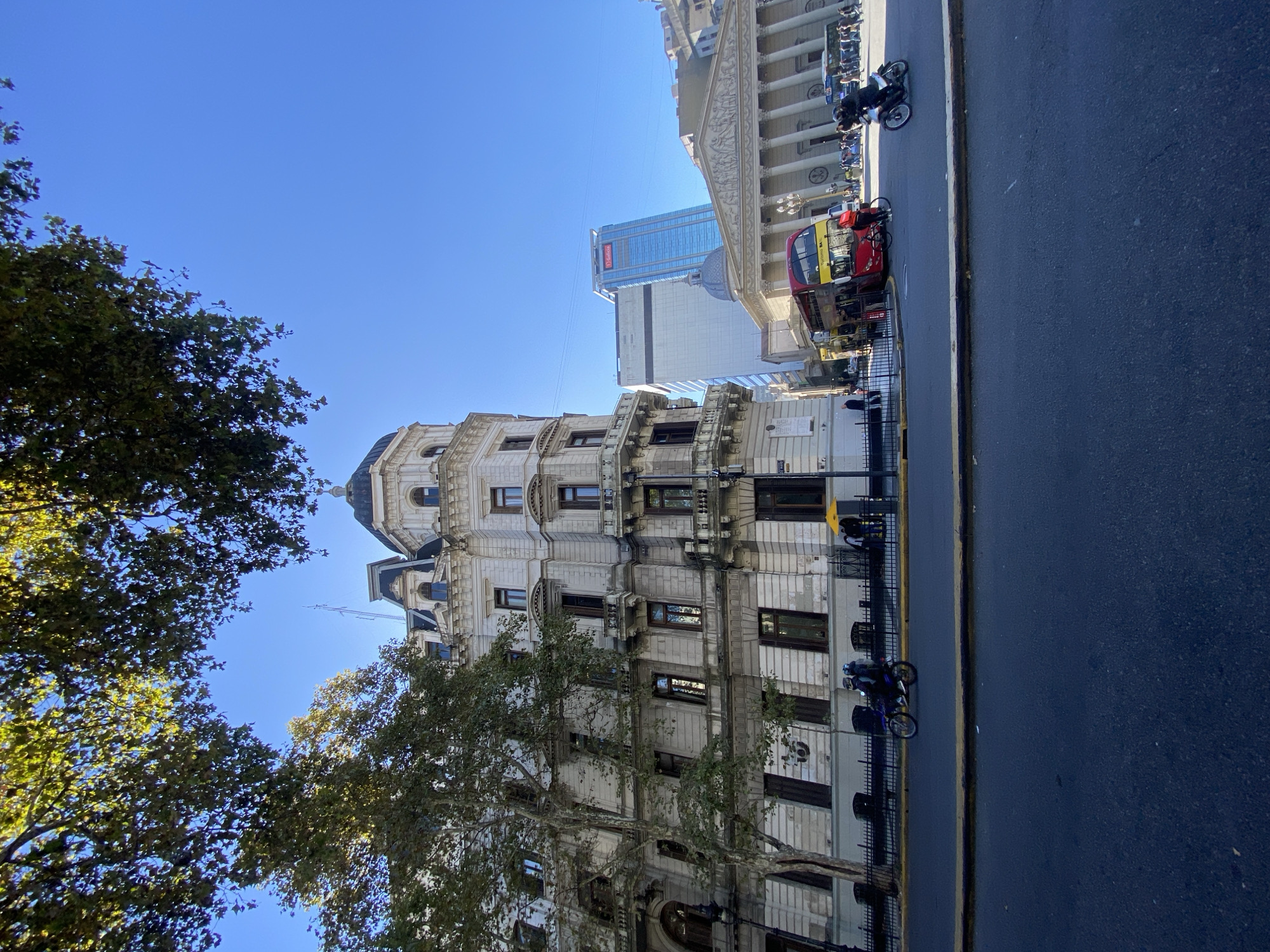 The image shows a street scene in Buenos Aires, Argentina, featuring several notable buildings. On the left is a detailed, light-colored building with multiple stories, dark-framed windows, and a domed top. Trees are partially obscuring the building's lower levels. In the background, to the right, is a larger, classical-style building with columns and a triangular pediment. A modern glass skyscraper rises behind these structures. The street is dark pavement with a light-colored curb line. There is a black iron fence separating the building on the left from the sidewalk. A double-decker tour bus and several motorcycles are parked along the street. The sky is clear and blue.