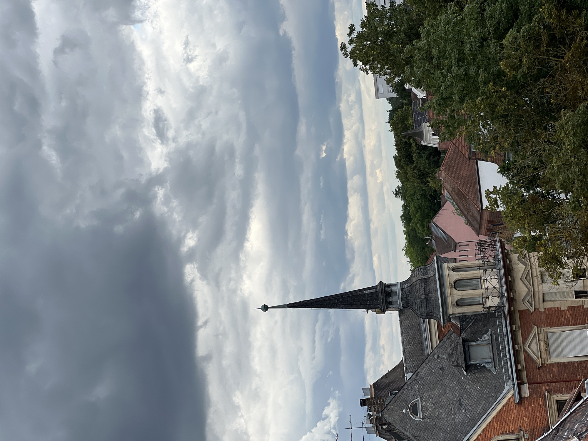 The image shows a view of rooftops and a spire in Straubing, Germany, under a cloudy sky. In the foreground, the corner of a red brick building with a gray tiled roof is visible. Above this building, a tall, dark, pointed spire with a small globe on top rises prominently. Further back, various other rooftops, including pink and white structures, are seen amidst lush green trees. A modern, light-colored building stands on the right side of the frame. The sky is filled with dense, dark, and lighter gray clouds, suggesting an overcast or possibly stormy day. There are no people or animals visible in the image. The scene captures a section of the urban landscape characterized by traditional architecture and natural elements.