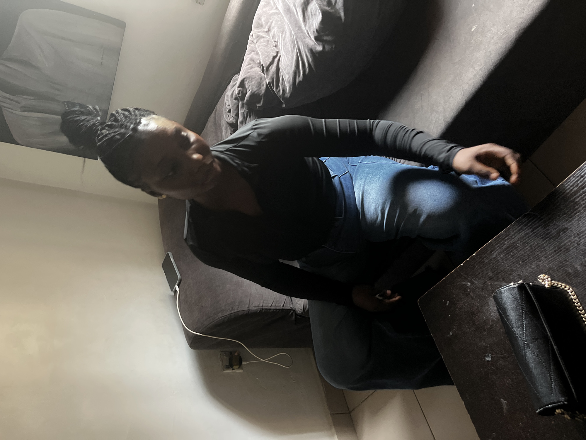 The image shows a woman with dark skin and braided hair in a bun, wearing a black long-sleeved shirt and blue jeans. She is sitting on a dark gray sofa in a dimly lit room. To her left, on a dark wooden table, is a black clutch purse with a gold chain. In the background, a large black and white framed artwork is mounted on the wall. A white cable runs from an electrical outlet on the wall to a smartphone on the sofa. The scene appears to be an indoor setting, potentially a living room, within a residence in Abuja, Nigeria.