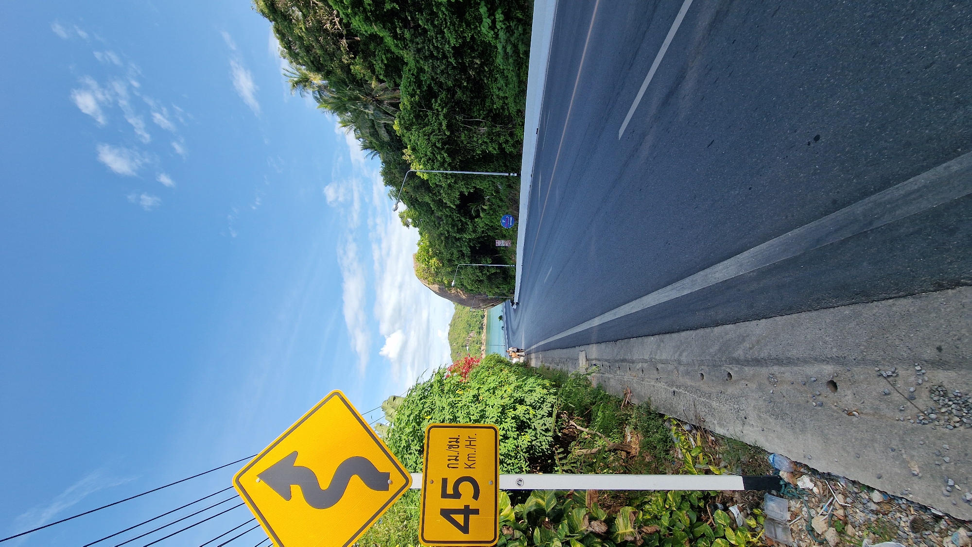 A straight, upward-angled, eye-level shot captures a coastal road in Baan Chaweng Noi, Thailand. The foreground features a yellow, diamond-shaped warning sign with a black, wavy arrow, indicating a winding road ahead. Below it, a rectangular sign displays "45" in black digits, followed by Thai text and "Km./Hr." in English, signifying a speed limit. A white pole supports the signs. To the left of the signs, a concrete sidewalk is visible, bordered by greenery and some debris. The road itself is asphalt with white lane markings, curving gently into the distance. Lush green trees and palm trees line the right side of the road, with a large rock formation embedded in the foliage. In the background, a body of water, likely the ocean, is visible, with a clear blue sky dotted with white clouds above. Two people and a scooter are seen walking along the sidewalk in the mid-ground. Streetlights are positioned along the roadside.