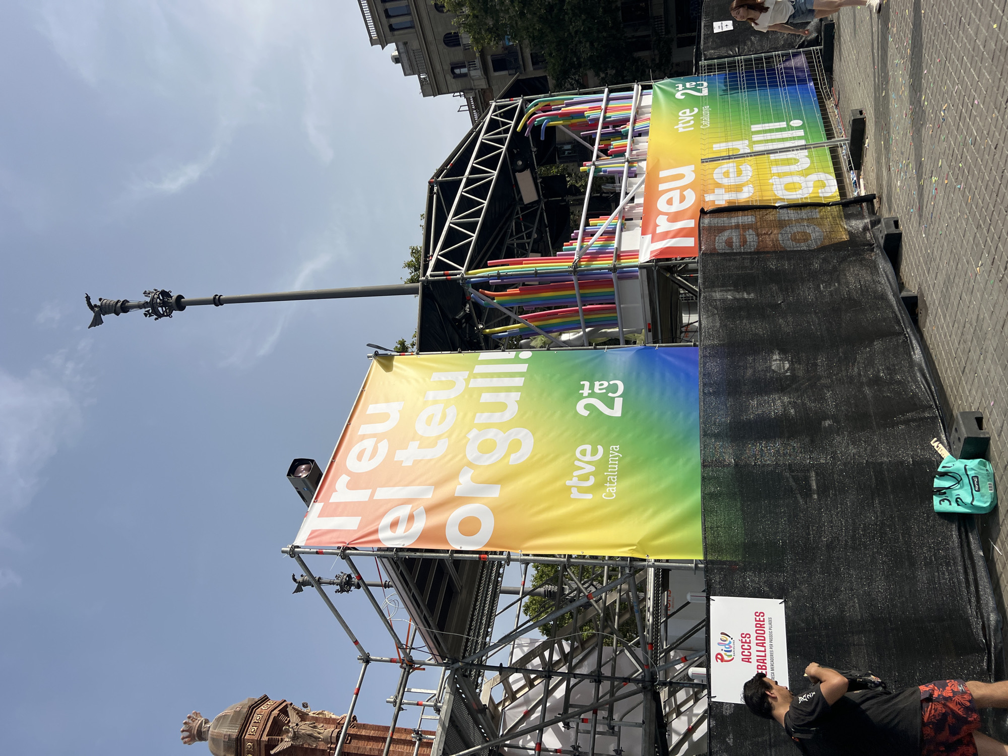 The image captures a vibrant outdoor scene in Barcelona, Spain, likely during a Pride event given the prominent rainbow banners and confetti on the ground. A large, scaffolding-supported stage is erected, adorned with colorful rainbow-themed decorations and banners that read "Treu el teu Orgull!" (Show your Pride!) with "rtve Catalunya 25" branding. To the left of the stage, a brick tower with a distinctive crown-like top and winged sculptures is visible, possibly a landmark within the city. A man in a black t-shirt and patterned shorts is standing near a white sign that says "Pride Barcelona Accés Ballarada", holding his phone. A woman in denim shorts and a white t-shirt is walking on the right side of the image, past a metal fence. The ground is paved with square tiles, and scattered confetti adds to the festive atmosphere. The sky is a clear blue with a few wispy clouds.