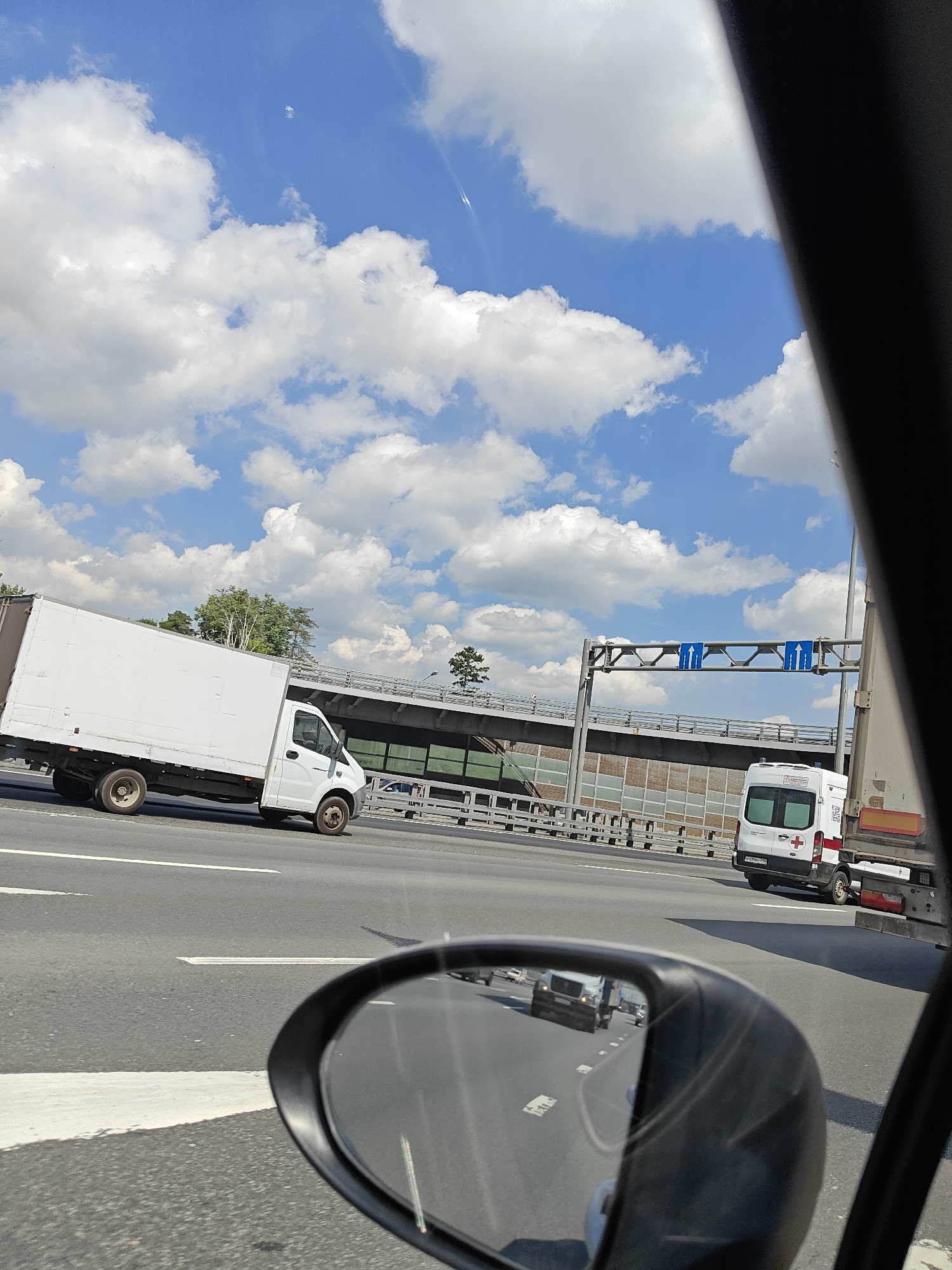 The image captures a view from inside a vehicle on a highway, looking towards the right side. In the foreground, a black rearview mirror reflects oncoming traffic, including a white truck and other vehicles. The road appears to be a multi-lane highway with clear white lane markings.
Further down the highway, a white box truck is driving in the left lane. To the right of the box truck, an overpass is visible, supported by metal structures and featuring sound barriers. Above the highway, blue road signs with white arrows indicating lane directions are mounted on a gantry.
In the right lane of the highway, a white ambulance with a red cross symbol on its rear is visible. This ambulance appears to be a medical vehicle. The sky above is blue with scattered white cumulus clouds, suggesting fair weather. In the distance, trees can be seen beyond the overpass.
The scene is consistent with a highway in an urban or suburban area. While the specific location of Mytishchi, Russia, is mentioned, there are no explicit visual cues within the image to confirm this location. However, the presence of a highway, overpass, and road signage are common in such environments.