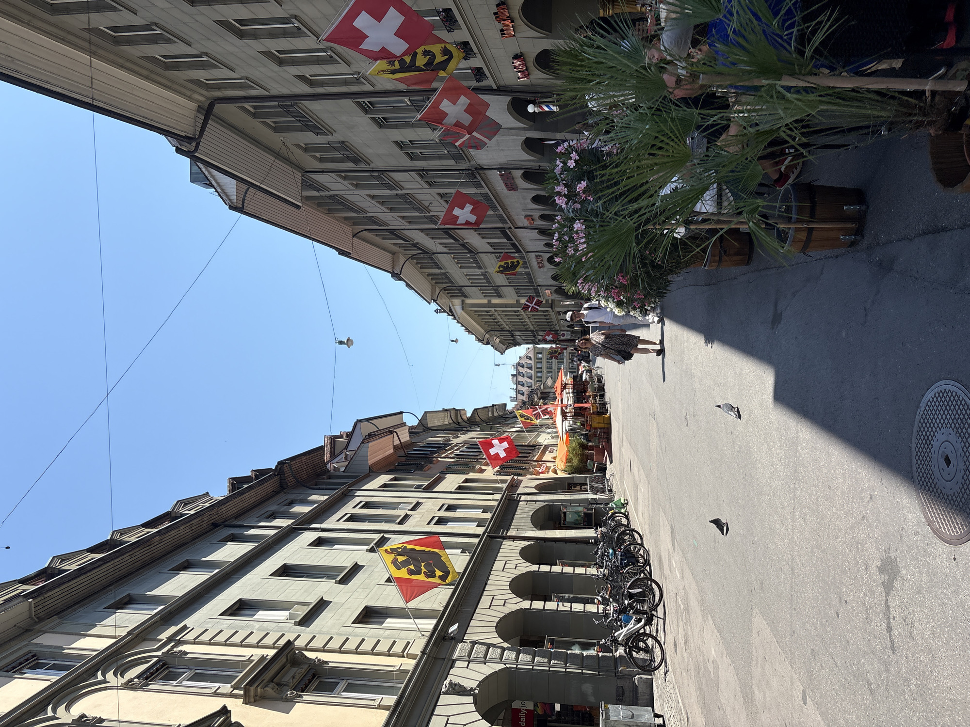 The image depicts Kramgasse in Bern, Switzerland, a narrow street lined with historic buildings on both sides. The architecture features arcaded ground floors, typical of Bern's old town. Several Swiss flags are prominently displayed, hanging from the buildings, alongside other flags, possibly regional or historical banners.
On the left side of the street, a row of parked bicycles is visible, suggesting a common mode of transport in the area. Further down the street, an outdoor cafe with tables and umbrellas is set up, with patrons seated and enjoying their time. Several potted plants, including palm trees and flowering bushes, add a touch of greenery and ambiance to the scene.
A few pigeons are seen on the paved street, and shadows of buildings and trees cast long shapes across the asphalt. The sky is clear and blue, indicating a sunny day. Two individuals are walking on the left side of the street, while other people are visible in the background, suggesting a lively and active urban environment. The overall impression is of a picturesque and well-preserved historic street in a European city.