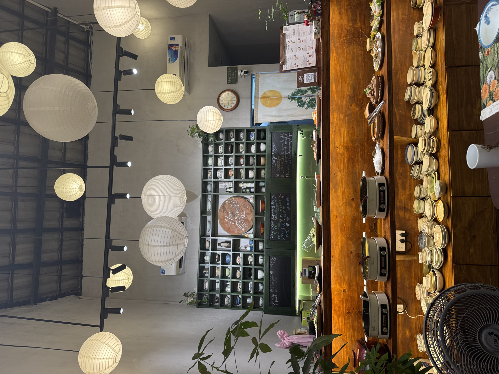 This image depicts the interior of a vegan buffet restaurant named "Hoa Gốm" in Đà Nẵng, Vietnam. The scene is illuminated by numerous white paper lanterns of varying sizes suspended from the ceiling, complemented by black track lighting fixtures. The walls are light gray, and the ceiling features exposed dark metal structural elements.
In the center, a multi-tiered wooden buffet counter is prominently displayed. The upper tier holds several serving dishes with prepared food items and three white electric chafing dishes, each containing food and labeled "Bear." Green LED strip lighting illuminates this tier. The lower tier of the buffet counter features extensive stacks of small, ceramic bowls and plates of various designs, intended for customer use, alongside additional prepared food. A black "ASIA" brand electric fan is visible on the floor in the immediate foreground, positioned in front of the lower buffet tier.
The back wall features a large, dark green shelving unit filled with numerous small, decorative ceramic bowls and cups. A circular wooden plaque with "Hoa Gốm" (Ceramic Flower) and floral designs is centered on this unit. Below the shelves, two black chalkboards display information. The right chalkboard details "Opening hours" (Mon-Sun: 5:00-9:00 PM; Day of the Lunar Month: 11:00 AM - 2:00 PM) and "Buffer Vegan" pricing at 100,000 VND per person, with a notice about prepayment. The left chalkboard mentions "V.I.P. Hoa Gốm."
To the right of the shelving unit, an "INVERTER" air conditioning unit is mounted high on the wall. Below it, an analog wall clock displays approximately 11:30. Next to the clock, a doorway is visible, covered by a light-colored fabric curtain depicting a yellow sun and green trees. Above this doorway, a sign reads "Nhà Vệ Sinh / Toilet," and a security camera is mounted below the sign. A wooden stand with a menu board for drinks, including a QR code, stands near the doorway. Small potted plants are strategically placed on the shelves and near the buffet counter.
In the bottom right foreground, a section of a table is visible, featuring a white mug, a blue-rimmed ceramic bowl, and a patterned placemat. No individuals are present in the image, and no actions or interactions are depicted.