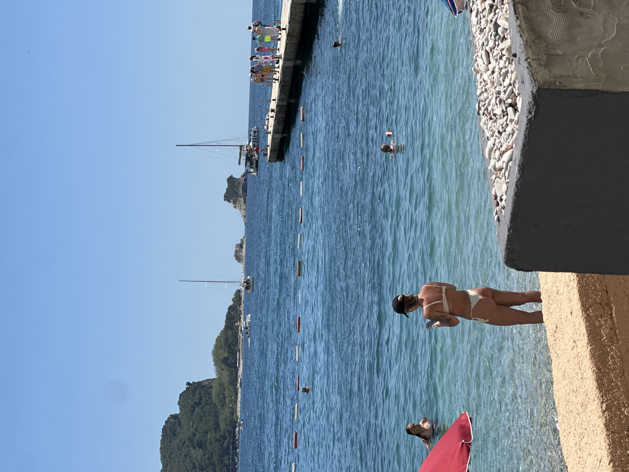 The image depicts a sunny day at a beach in Budva, Montenegro. Several people are visible, some standing on a pier and others swimming in the clear blue water. A sailboat is anchored in the background, and lush green hills rise behind the coastline. The sun is high in the sky, suggesting it's midday. The water is calm and inviting, with a few buoys marking a swimming area. The overall atmosphere is relaxed and recreational, with people enjoying a typical summer day at the seaside.