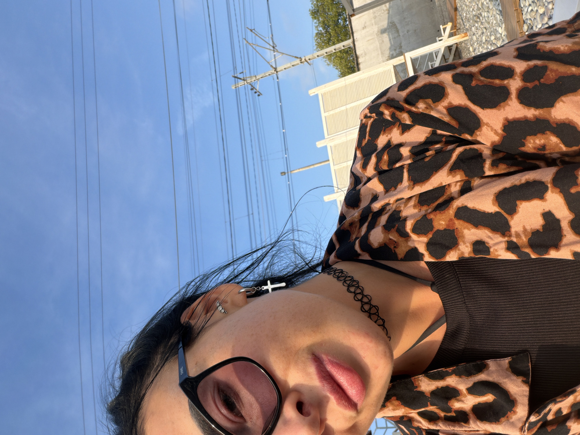 A close-up, outdoor, eye-level, medium selfie features a woman wearing a leopard print jacket, a black ribbed tank top, a black choker necklace, a silver hoop earring with a cross pendant, and black-framed glasses. She has dark hair and fair skin with freckles. The background includes a clear blue sky with power lines, a utility pole, and a light-colored wooden structure. The lighting suggests it is either morning or late afternoon.