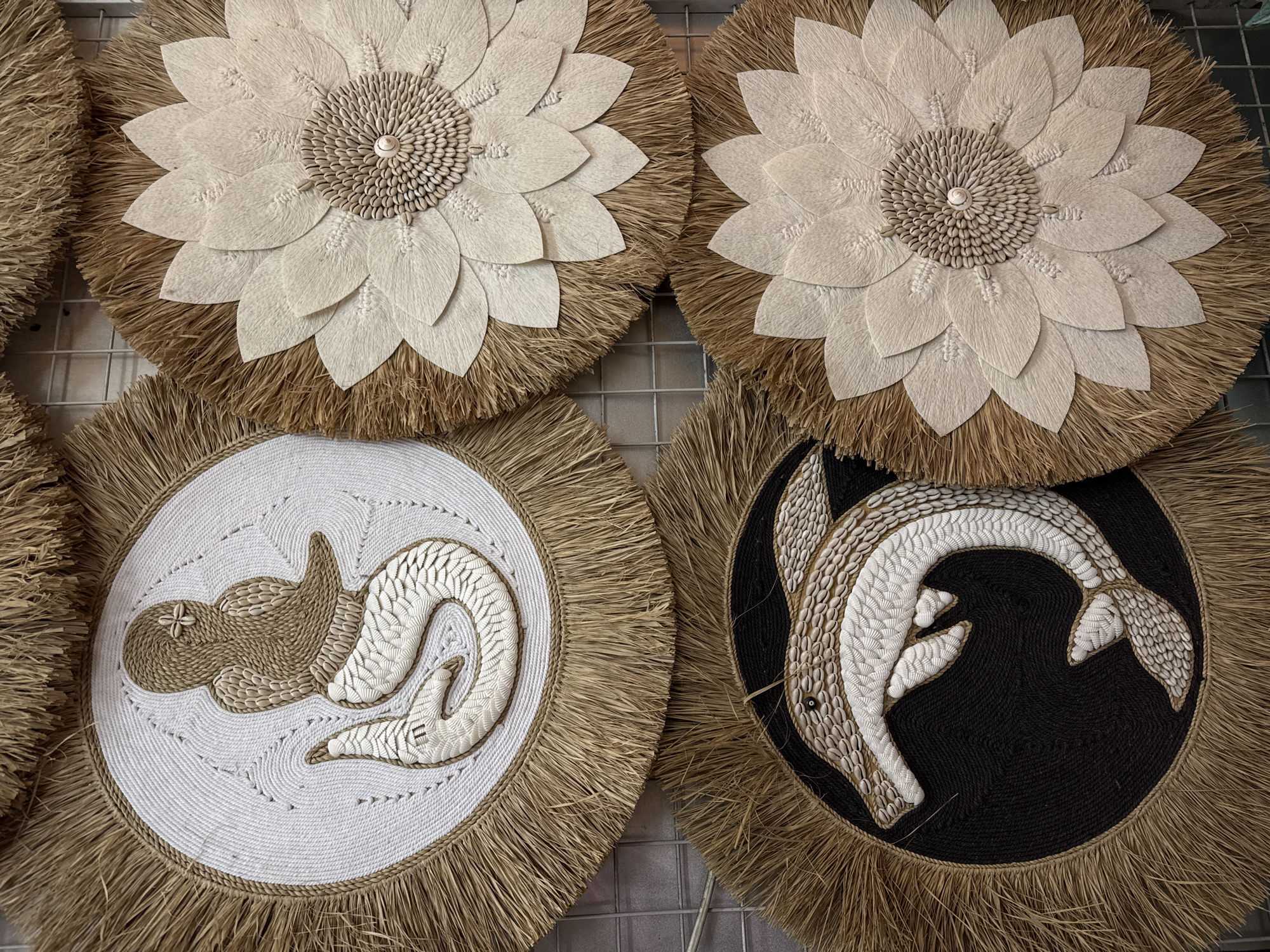The image displays four circular wall hangings arranged in a two-by-two grid, likely presented for sale in Kuta, Indonesia. The hangings are made of natural materials, primarily straw or raffia for the fringed outer edges and woven or fibrous material for the inner circular designs.
The top-left hanging features a stylized mermaid figure crafted from beige and white shells and rope, set against a white woven background. The mermaid is depicted with a shell detail on its head.
The top-right and bottom-right hangings are identical, featuring a floral design composed of layered, light-colored petal shapes made of a fibrous material. The center of each flower is a dense arrangement of small shells, with a single larger shell at the very center. The straw fringe surrounds these floral designs.
The bottom-left hanging showcases a dolphin figure, also constructed from beige and white shells and rope, against a solid black woven background. The dolphin is depicted in mid-leap.
The hangings are mounted on a metal grid structure, common in market stalls or retail environments. The lighting suggests an indoor or shaded outdoor setting. There are no people or animals present in the image. The overall aesthetic is artisanal and beach-themed.