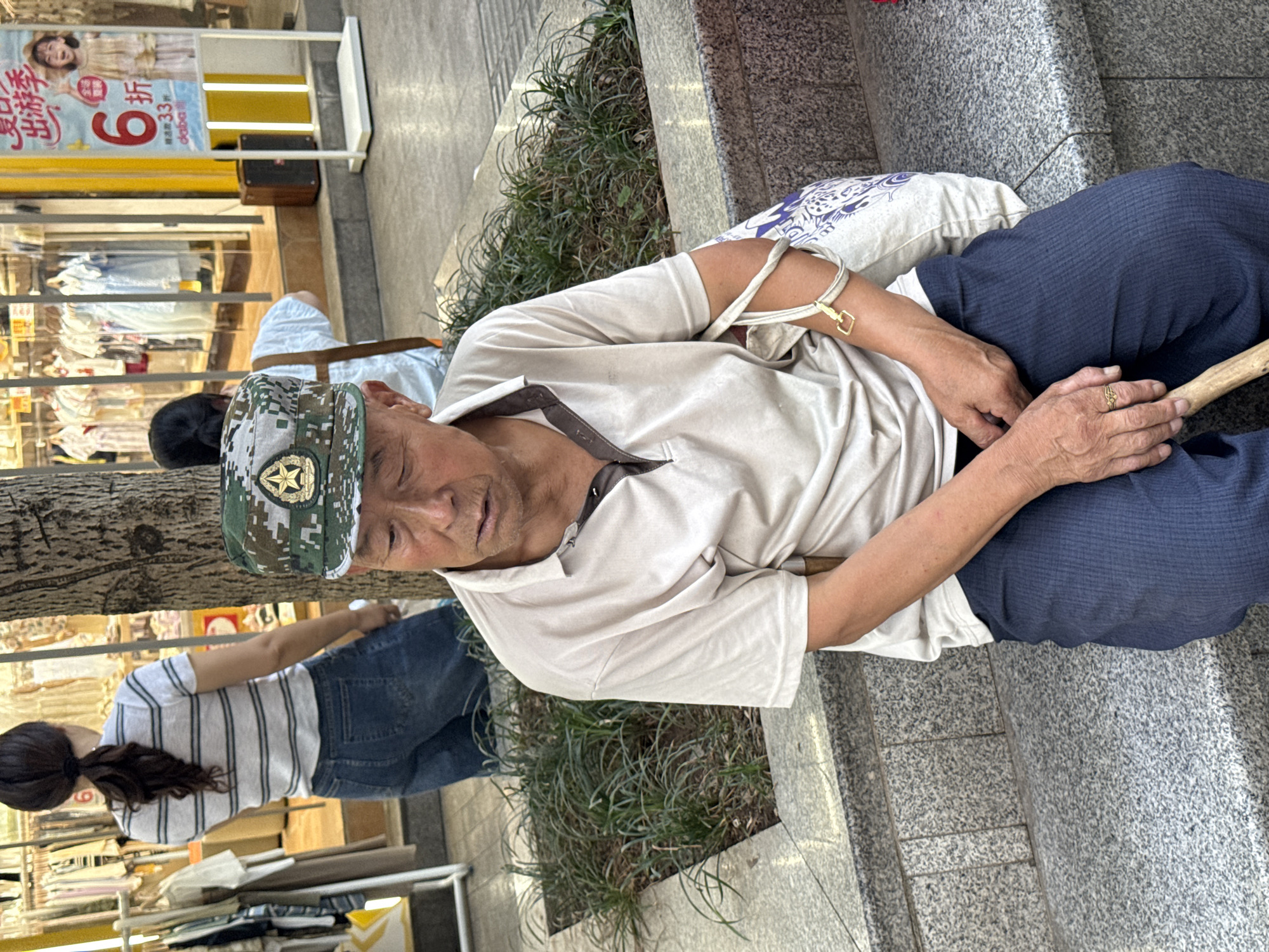 The image depicts an elderly man resting on a granite-patterned stone ledge in what appears to be an urban commercial area or street in Jiangjin, China, during daytime. The man is wearing a light beige short-sleeved polo shirt with a dark collar, dark blue pleated pants, and a camouflage military-style cap featuring a prominent golden star emblem. He is holding a light-colored wooden walking stick with his hands clasped in his lap. A white pillow with blue, patterned designs is placed behind him for comfort. A gold-colored ring is visible on his left ring finger, and a distinctive golden rectangular buckle is fastened to a strap around his right upper arm. His eyes are partially closed, suggesting he is either sleeping or deeply resting, with a seemingly weary expression.
To his right, there's a planter box containing dark green, spiky foliage. In the background, behind and slightly to the left of the resting man, another person is seen from the back, wearing a striped t-shirt and jeans. Beyond them, a shop window displays various items of clothing. A prominent advertising banner is visible in the shop window, featuring a smiling woman in a light-colored dress. The banner contains Chinese text that reads: "夏日出游季" (Summer Travel Season), "6折" (60% off or 40% discount), "清凉透气 33元起" (Cool and breathable, starting from 33 yuan), and the brand name "debao". A dark brown box, possibly a speaker, sits on the ground near the advertisement. A tree trunk partially frames the left side of the scene. The ground is paved, indicating a public sidewalk or plaza. The overall scene suggests a moment of rest amidst a busy urban environment.