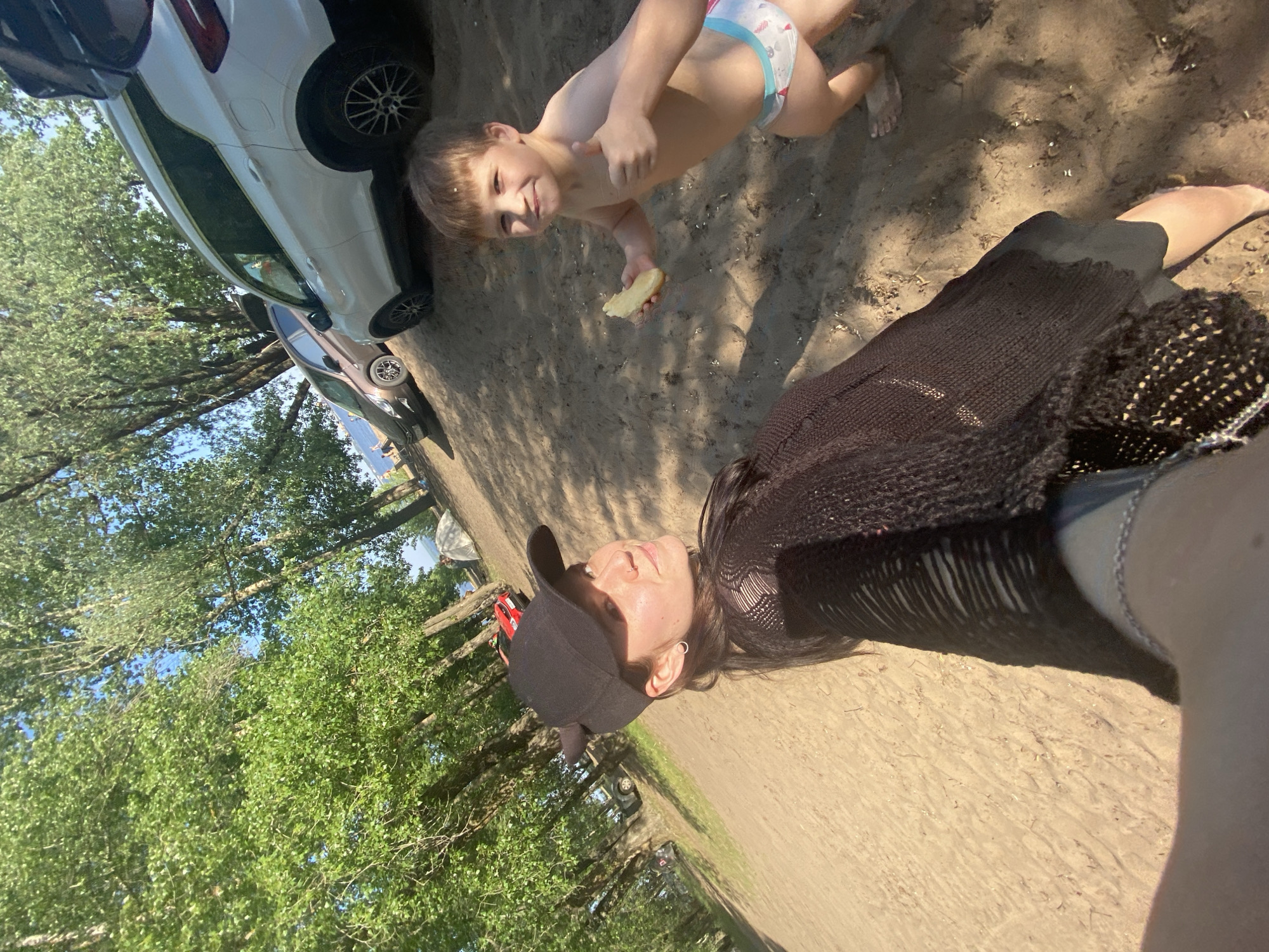 The image shows a sunny outdoor scene, possibly in a park or recreational area near trees, in the city of Chaikovsky, Russia. A young boy, wearing underwear, is seen from a high angle holding what appears to be a piece of bread or cake. A person, likely a woman, wearing a black top, jeans, and a black cap is in the foreground, suggesting she is holding the camera. Several parked cars are visible in the background. The ground is covered in sand.