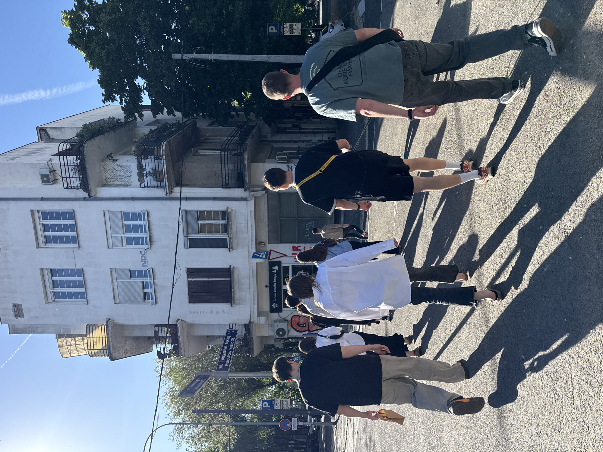 The image shows a group of people walking on a street in Belgrade, Serbia. The location is identified by a street sign indicating "ul. Gundulićev venac," which translates to Gundulićev venac Street. There are a total of six people in the group. The street is paved, with buildings visible in the background. The buildings appear to be residential or commercial, and there are trees that provide shade. There is a sign on one of the buildings reading "Užička komplet lepinja" and "NDC". Street signs and parking signs are also visible. The sunlight casts long shadows, suggesting it is early morning or late afternoon.