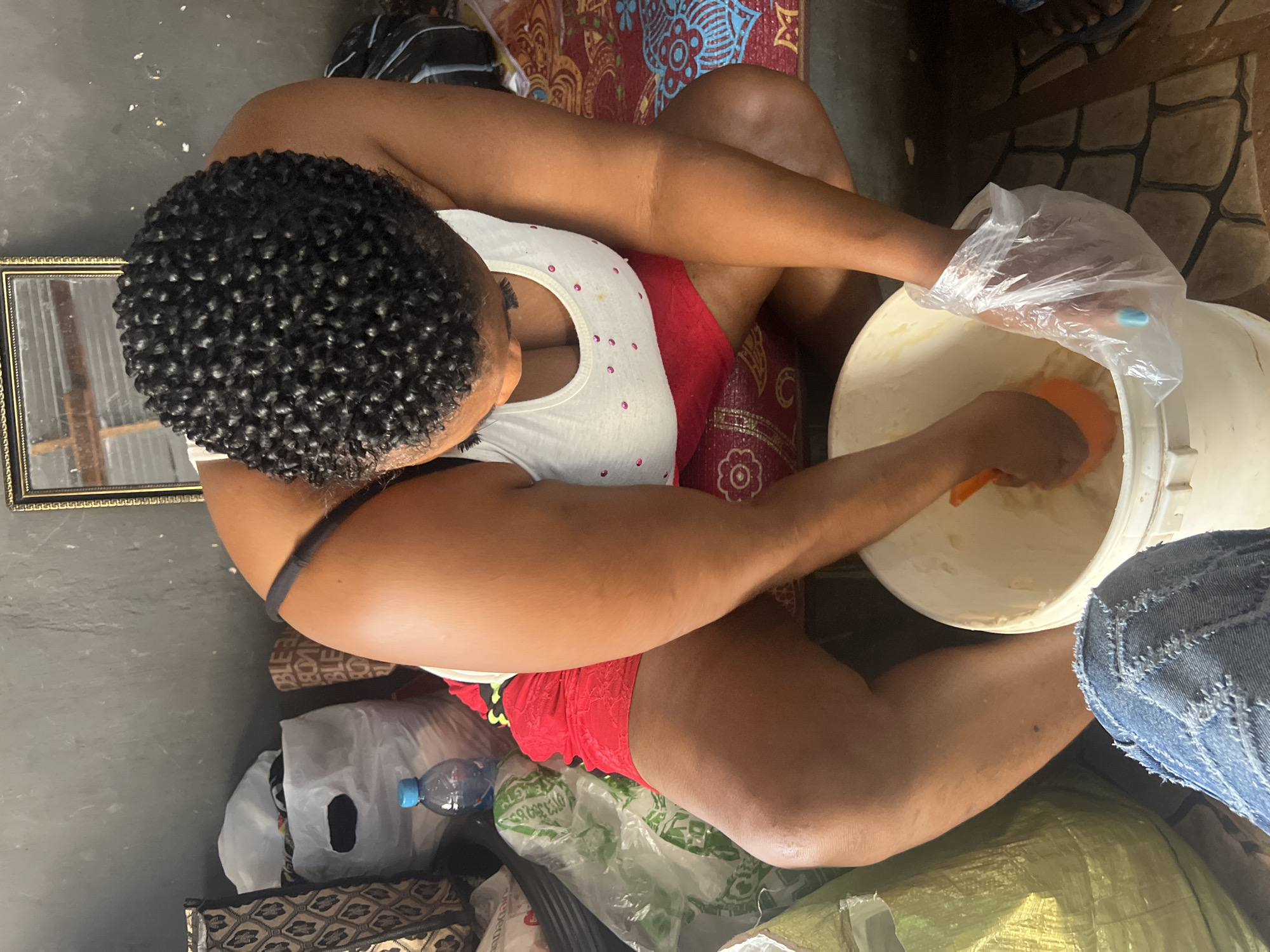 In a dimly lit room in Nkpor, Nigeria, a Black woman with tightly coiled hair, wearing a white tank top adorned with pink polka dots and red shorts, kneels by a large white bucket. Her arms are submerged in the bucket's contents, which appear to be a thick, pale substance, possibly dough or a mixture for soap making. She wears a clear plastic glove on her right hand and holds an orange scooper. The floor is tiled, and various bags and personal belongings are scattered around the room, including a partially visible blue water bottle and a framed mirror on the wall. The overall atmosphere suggests a domestic setting where a task involving manual labor is underway.