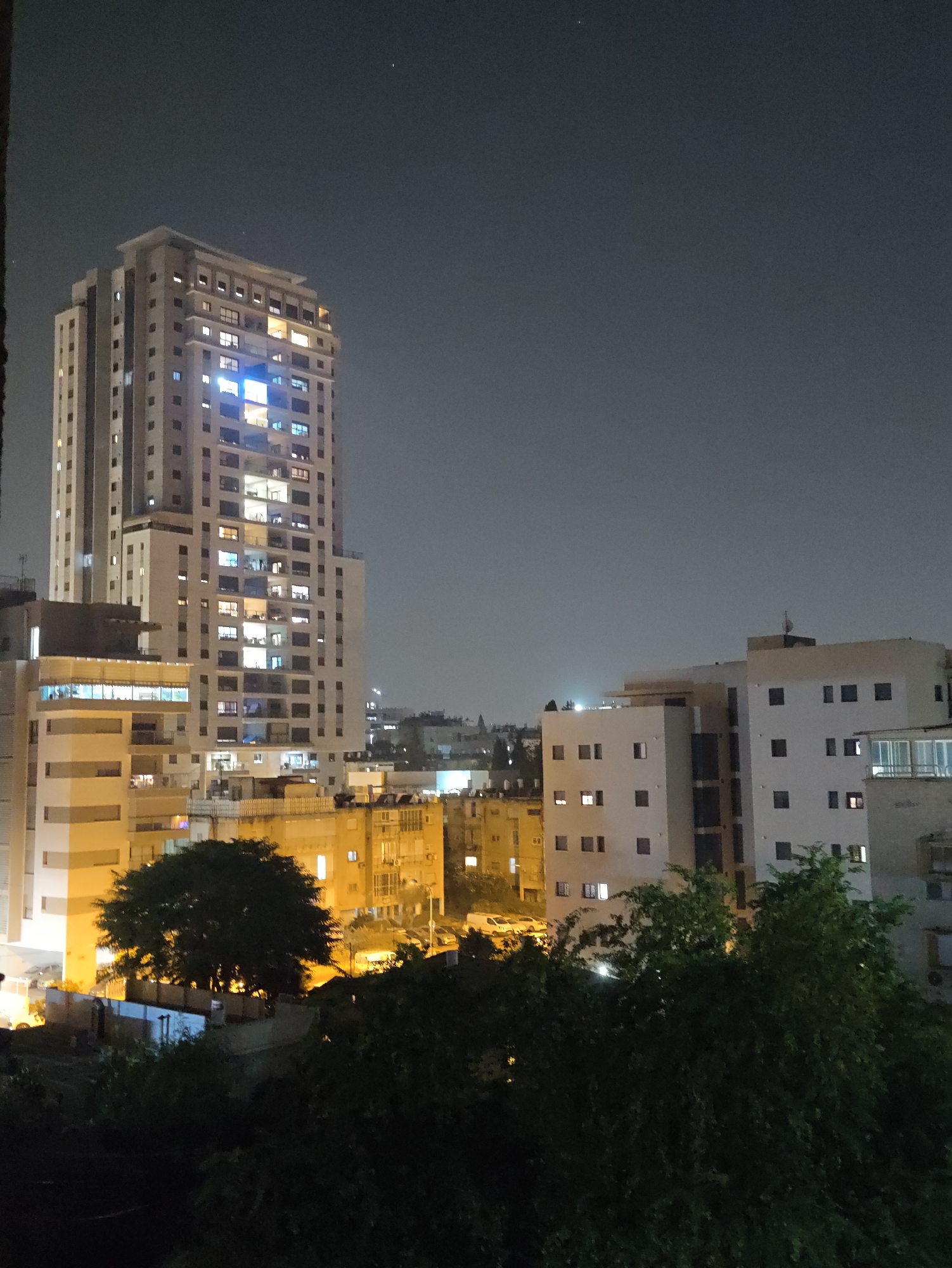 The image depicts a nighttime cityscape with several buildings illuminated by interior lights. In the foreground, a large, dark-leaved tree dominates the lower portion of the frame, obscuring some of the lower structures. Behind the tree, a street is visible with parked cars.
The focal point is a tall, light-colored apartment building with many windows, most of which are lit, suggesting occupancy. This building has a modern, rectangular design. To the right of this building, a slightly shorter, also light-colored building with a flat roof is visible, featuring multiple windows, some of which are illuminated. Further to the right, another building with a balcony or terrace is partially visible. In the background, other structures and lights indicate a dense urban environment. The sky is dark and appears overcast or hazy, with a faint light or star visible in the upper right quadrant. The overall scene conveys a sense of urban life at night. The buildings and tree suggest a residential area within a city. This view is consistent with urban landscapes in Petah Tikva, Israel, known for its mix of modern and older residential buildings.