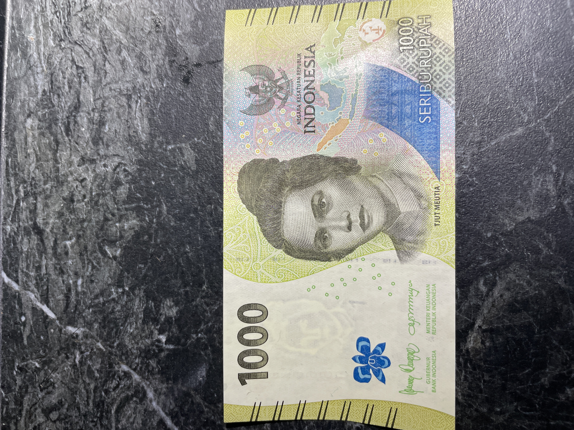 The image shows a 1000 Indonesian Rupiah banknote lying on a dark, textured surface. The banknote features a portrait of Tjut Meutia, national coat of arms of Indonesia, and the denominations "1000" and "SERIBU RUPIAH". The text "NEGARA KESATUAN REPUBLIK INDONESIA" and the signatures "GUBERNUR BANK INDONESIA" and "MENTERI KEUANGAN REPUBLIK INDONESIA" are visible.