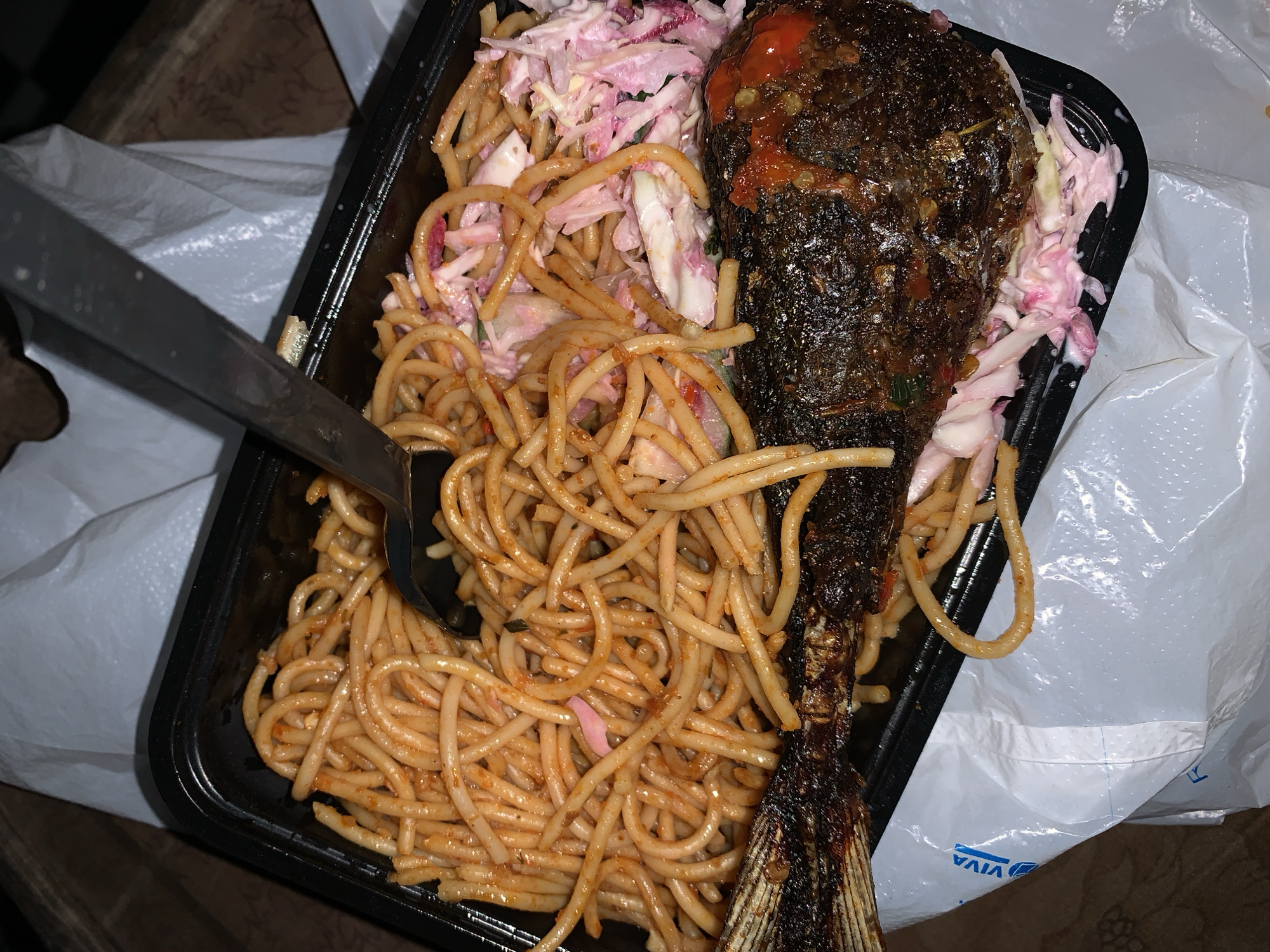 An overhead shot captures a takeaway meal presented in a black rectangular plastic container, likely resting on a surface and partially on a white plastic bag. The meal consists of three main components: a whole cooked fish, a generous portion of spaghetti, and a side of coleslaw.
The fish, dark brown to black in color with some reddish seasoning or sauce visible, is positioned along the right side of the container, extending nearly its full length. Its skin appears crispy, and the tail fin is visible. To the left of the fish and occupying the majority of the container is a mound of spaghetti, featuring thin, curly strands coated in a light orange or reddish sauce. Nestled between the fish and the spaghetti is a portion of coleslaw, made from shredded white and red/purple cabbage mixed in a creamy dressing.
A metal spork (spoon-fork hybrid) is partially visible on the left side of the container, resting in the spaghetti, suggesting the meal is either about to be eaten or is in the process of being consumed. The white plastic bag underneath the container has some blue text, with "VIVA" clearly discernible, though appearing upside down. The general presentation indicates a casual, ready-to-eat meal, typical of a takeaway service in a setting like Rafin Par, Nigeria. No specific time of day or weather conditions can be inferred from the image, nor are any emotions conveyed.