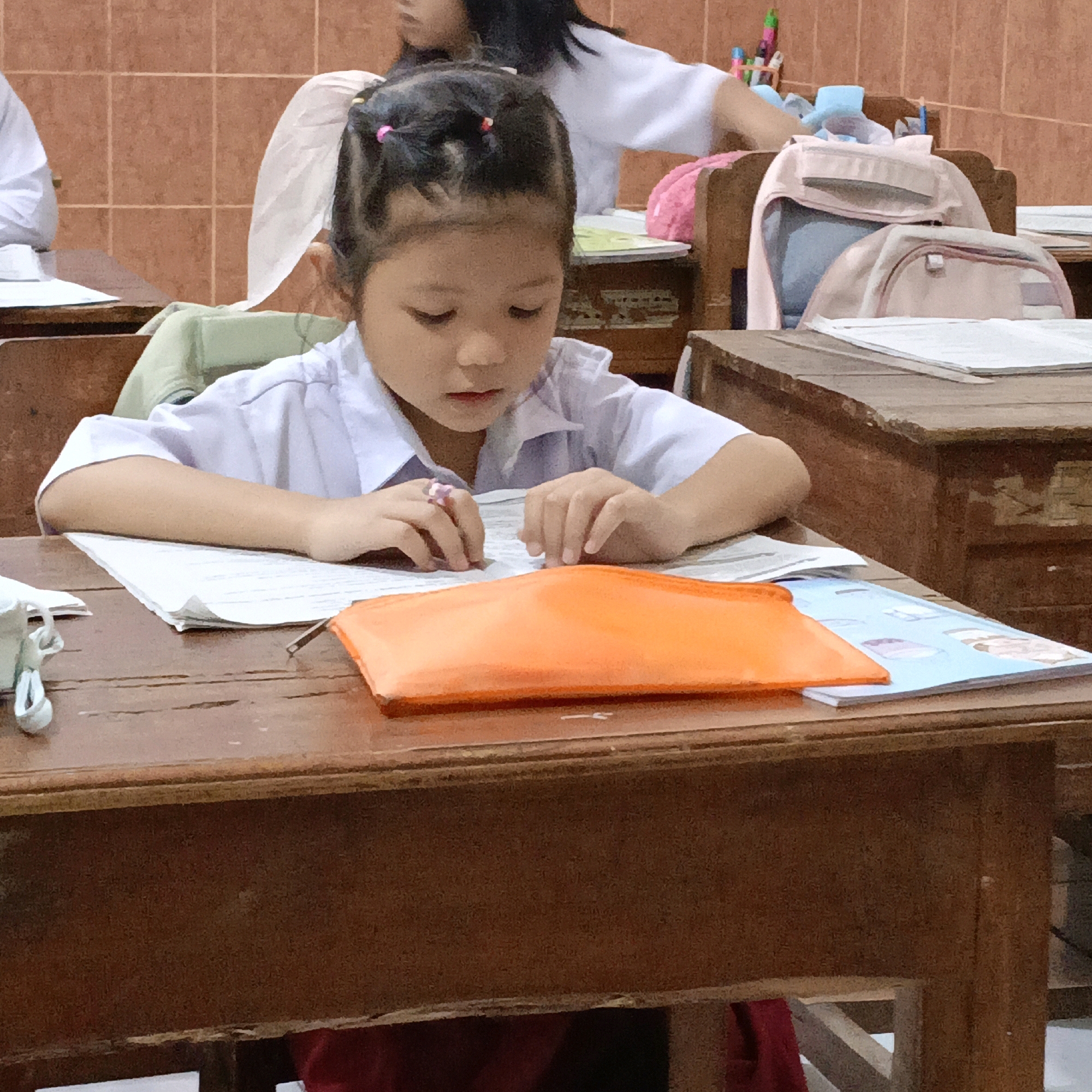 The image depicts a young girl, appearing to be of East Asian descent, seated at a wooden desk in what appears to be a classroom setting. She is wearing a white school uniform shirt and has her hair styled in two pigtails with pink elastics. She is looking down at papers on the desk and her hands are resting on them. One hand is wearing a ring with a pink stone. In front of her is an orange pencil case. Behind her, another student in a white shirt is partially visible. The background consists of tiled walls and wooden furniture, including backpacks on chairs. The overall impression is that of a school environment. The specific location in Salatiga, Indonesia, cannot be determined from the visual information provided in the image.