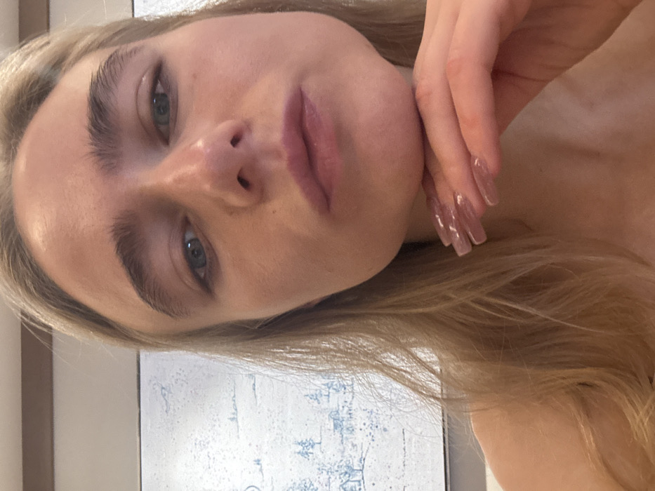 A fair-skinned woman gazes at the camera with a slight tilt of her head. She has light blonde hair and full lips. Her manicured nails are visible as her hand gently touches her face. Her eyebrows are thick and well-defined. The background shows a white wall with a glimpse of a framed picture featuring light blue drawings.
