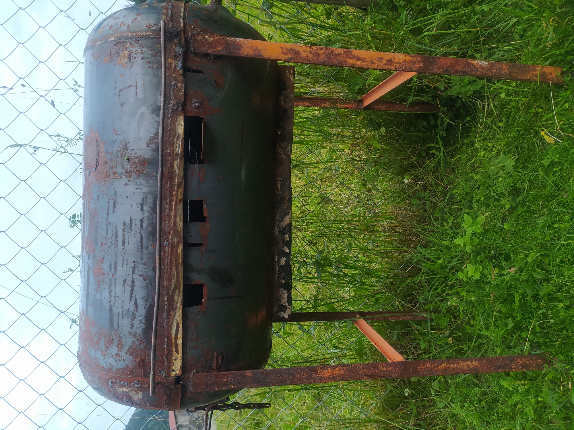 An old, rusty, cylindrical grill sits on four legs in a grassy area. The grill is made from a metal barrel, painted dark green and heavily weathered with patches of rust. It has a grate across the top, and several rectangular openings along its sides. The legs of the grill are orange, also showing signs of rust, and are connected by a horizontal support. The grill is positioned in front of a chain-link fence, beyond which is a faint view of trees and a building. The ground is covered in lush green grass and various small plants. The sky above is overcast. This scene appears to be in an outdoor, possibly rural, setting.
