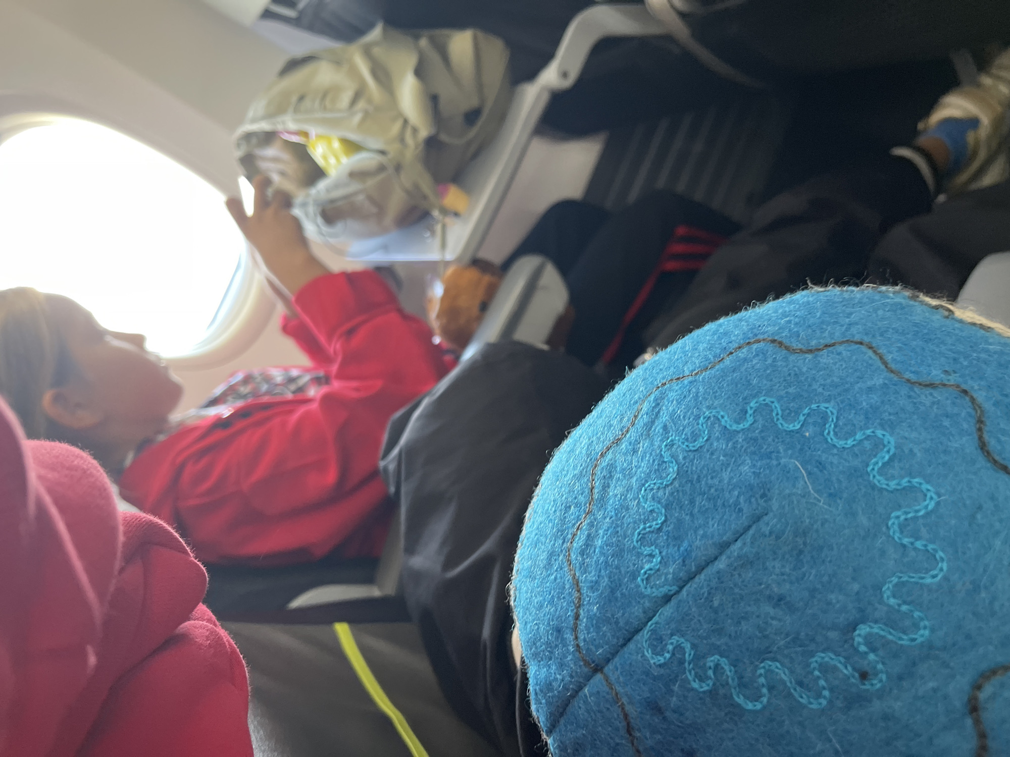 The image shows an interior view of an airplane cabin. In the foreground, a blue cap with decorative stitching is partially visible. Behind it, a person in a red jacket is seated, looking down at something in their hands and is near a tray table. Another person, partially obscured, is seated next to them. The environment suggests travel, specifically on an aircraft. The location of Choktal, Kyrgyzstan is not depicted in the image.