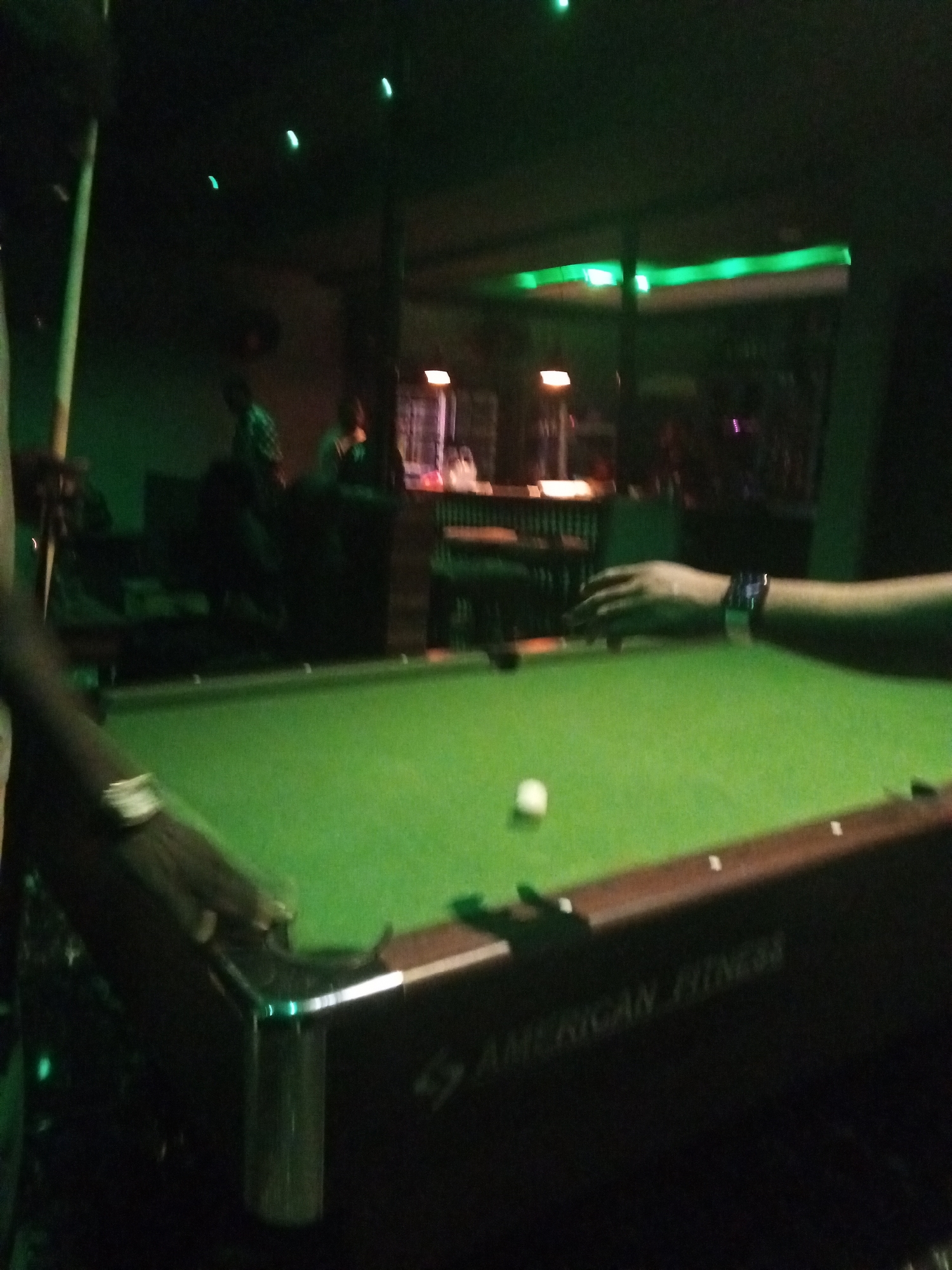 The image depicts a dimly lit indoor scene, likely a bar or recreational area, with a green pool table in the foreground. A white cue ball rests on the table. A person's arm and hand, wearing a watch on the wrist, are visible to the right, seemingly in the process of playing pool. Another person's arm and a pool cue are visible on the left side of the frame, also suggesting active gameplay. The background is dark and out of focus, with hints of people and furniture, including what appears to be a bar counter. Green ambient lighting is present, particularly along the ceiling and in reflections. The overall impression is one of casual recreation in a bar setting. Without additional context or visual cues, the specific location within Lokoja, Nigeria, cannot be definitively determined from the image alone. However, the style of the pool table and the presence of the "American Fitness" branding suggest it could be a commercial establishment.