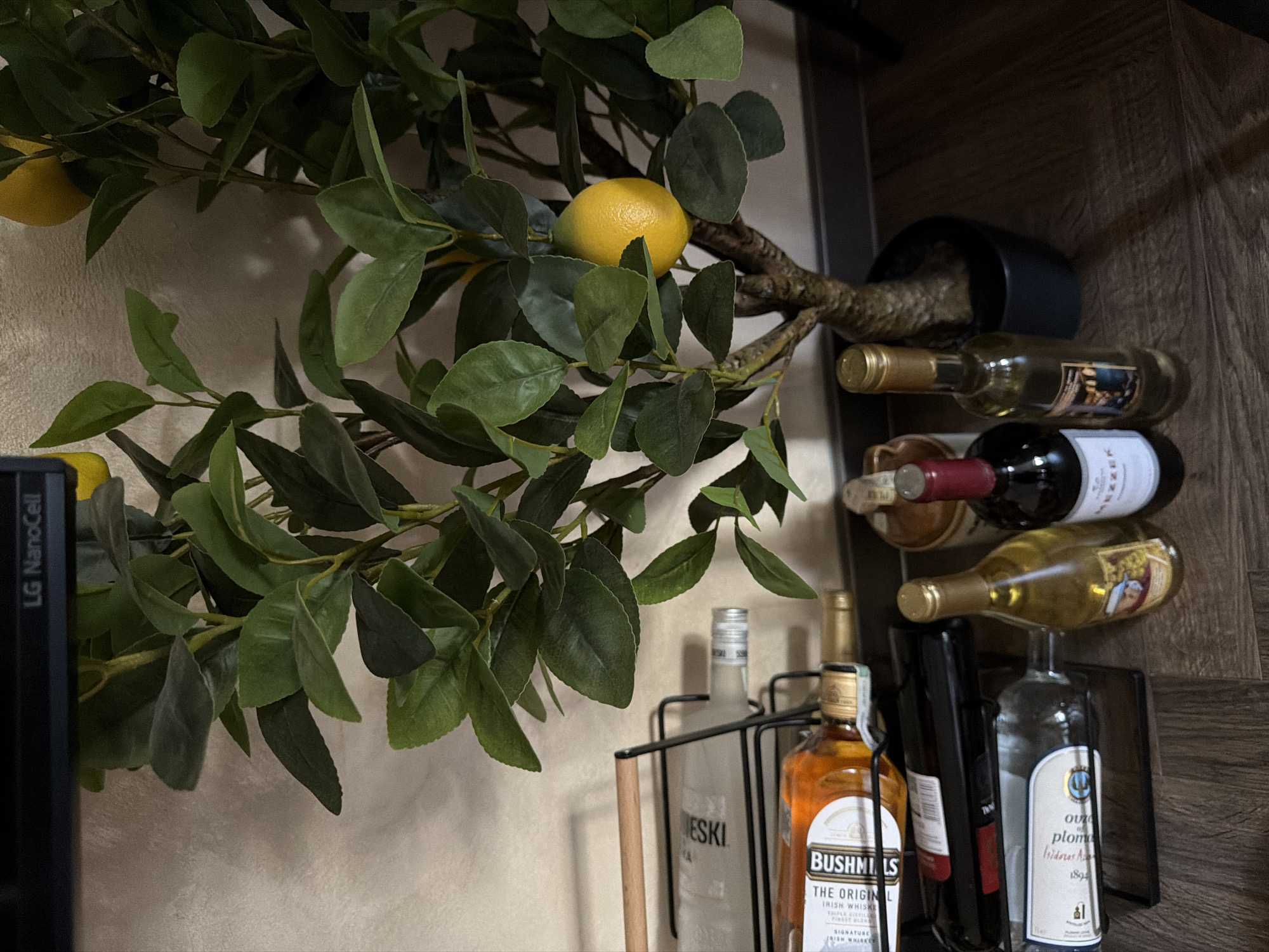 The image depicts an indoor scene in Sofia, Bulgaria, featuring a still life arrangement of alcoholic beverages and an artificial plant. A black metal wine rack is positioned on the left, holding three visible bottles. The top bottle is clear with a partially visible label including "ESKI". The middle bottle contains an amber liquid, labeled "BUSHMILLS THE ORIGINAL IRISH WHISKEY". The bottom bottle is clear with a partially legible label that includes "plom" and "1892". Behind the bottom clear bottle, a red-labeled bottle is partially obscured. To the right of the wine rack, a faux lemon tree with multiple green leaves and two bright yellow artificial lemons stands in a dark pot. The tree's trunk is visible. On the floor to the right of the wine rack and in front of the lemon tree pot, three additional bottles are present. The leftmost is a red wine bottle with "MEZZEK" visible on its label. The middle bottle is clear with a gold cap and a light-colored liquid. The rightmost bottle is partially visible, appearing to be a stoneware vessel with a red cap. The background consists of a light-colored, subtly textured wall. The floor is dark wood-grain. A black surface, possibly a television screen or monitor, bearing the text "LG NanoCell", is visible at the very top of the frame. No individuals are present in the image.