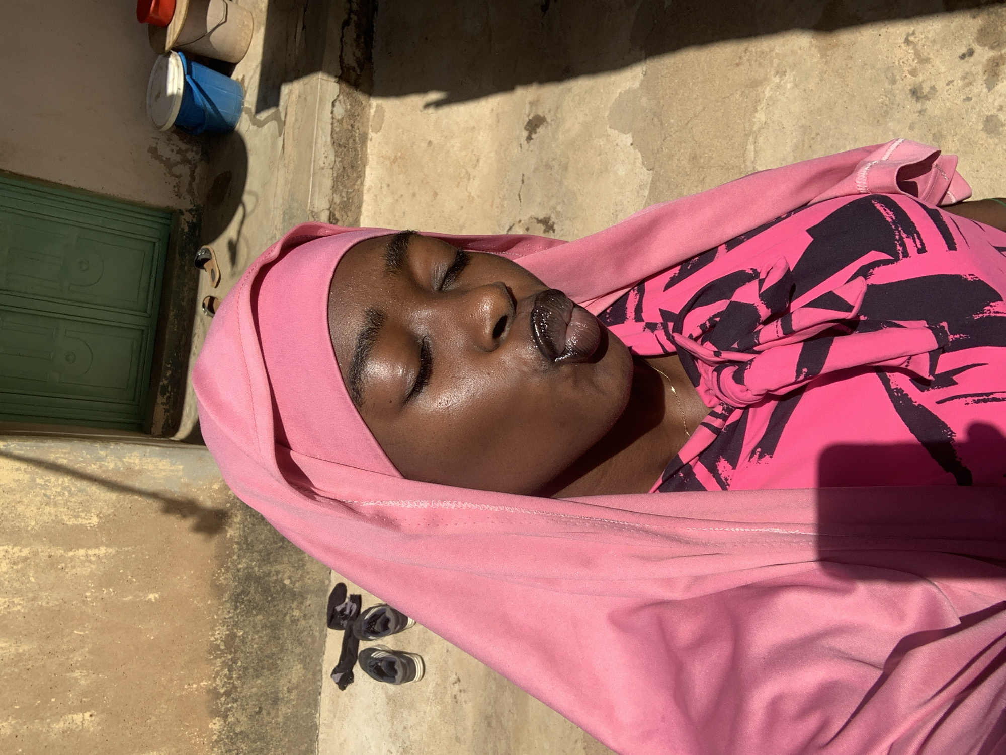 A woman is lying on a concrete surface next to a building in Rafin Par, Nigeria. She is wearing a pink headscarf and a pink patterned shirt. Her eyes are closed and her lips are puckered. A pair of shoes and a sock are on the ground next to her. A green door and several buckets are visible in the background. The scene is outdoors, illuminated by sunlight.