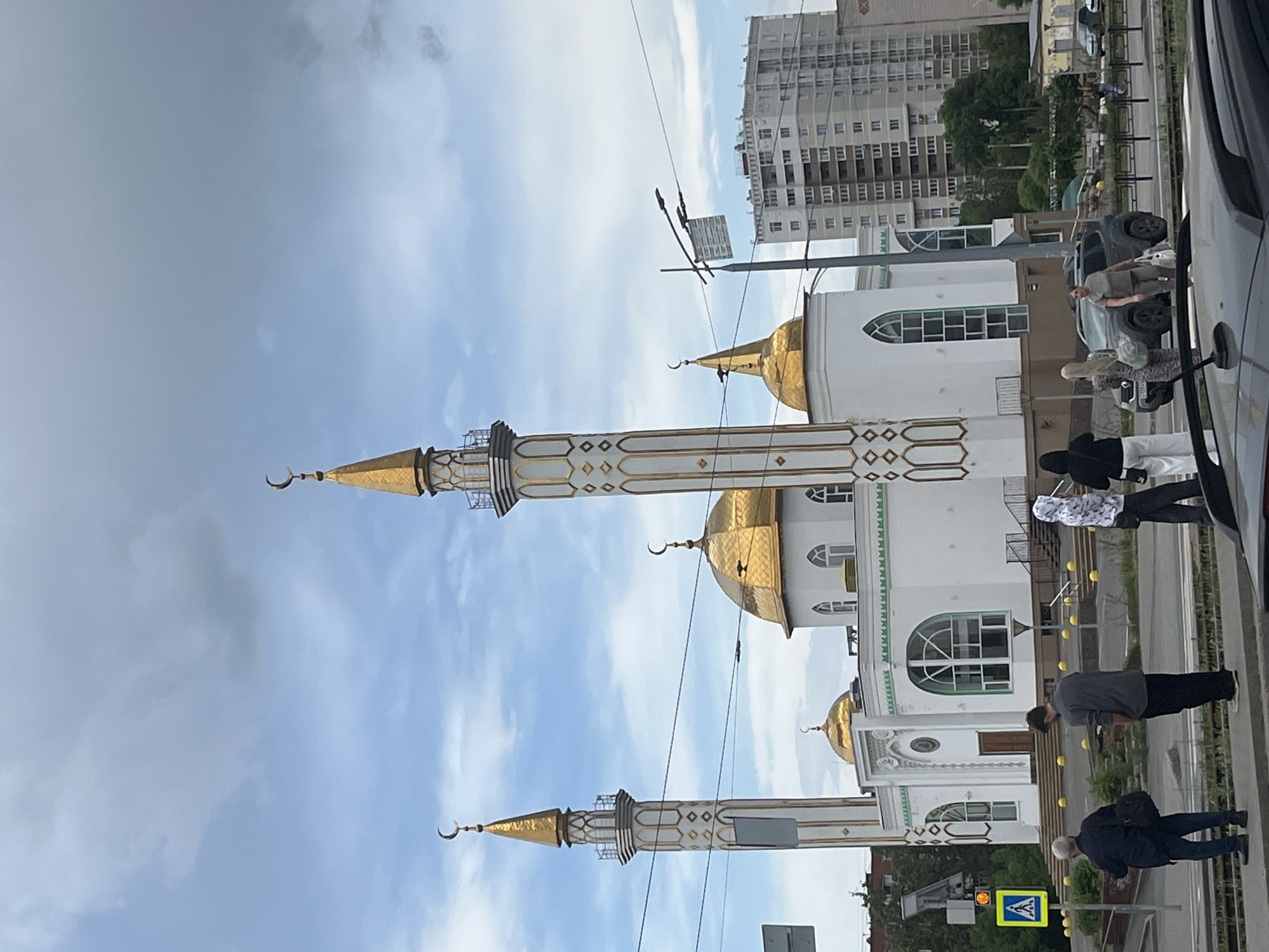 The image captures the Izhevsk Sobornaya Mosque against a partly cloudy sky. The mosque, primarily white with gold accents on its domes and minarets, stands on what appears to be a city street with pedestrians walking by. In the background, a tall, multi-story building is visible. The scene suggests an urban environment, with the mosque serving as a prominent architectural feature. Several pedestrians are walking past. A car is parked.