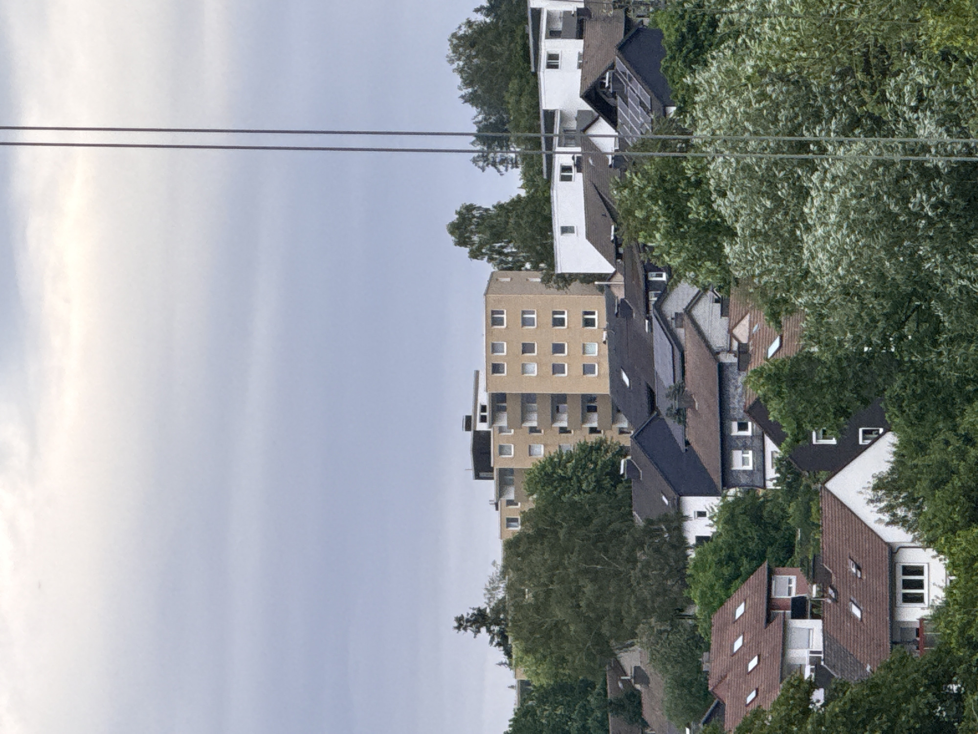 The image shows a view of residential buildings in Bad Berleburg, Germany. In the background, a tall, beige apartment building with a dark roof is visible, featuring multiple floors with evenly spaced windows. In front of this, a row of smaller, white and dark-roofed buildings are situated. Closer to the foreground, several houses with pitched roofs, some of which are covered with solar panels, are nestled among lush green trees. The sky is overcast and appears pale blue and white. Two thin, vertical metallic poles run through the right side of the frame, suggesting the presence of utility lines or scaffolding. There are no people visible in the image.