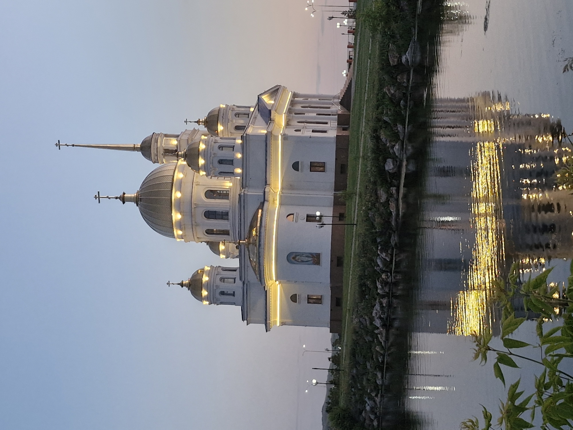 The image depicts the Assumption Cathedral in Kokshetau, Kazakhstan, at dusk. The white, neoclassical cathedral is illuminated by warm yellow lights along its roofline and around its windows, highlighting its golden domes and cross-topped spires. The building features large columns at its entrance and a mural on its side. In the foreground, a calm body of water reflects the illuminated cathedral, creating a shimmering visual effect. The shoreline is lined with grass, rocks, and a few sparse trees. In the background, hills are visible against a pale, twilight sky transitioning from blue to soft pink. Several lampposts are also present along the embankment. No people are visible in the image.