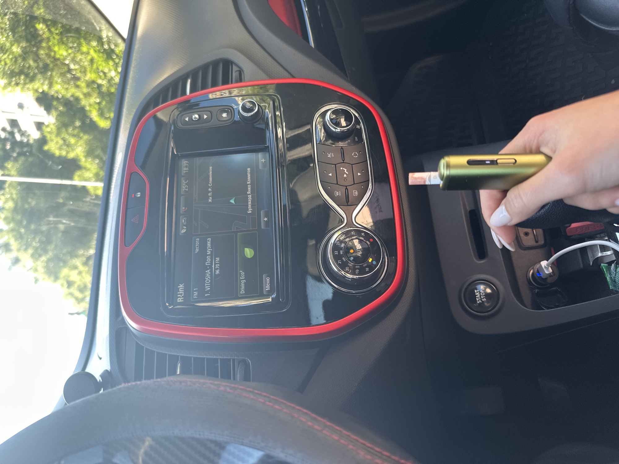 The image shows the interior of a car, likely parked. In the foreground, a person's hand is holding a gold-colored electronic device with a beige cigarette-like object inserted. The car's dashboard is visible, featuring a central infotainment screen displaying "R-Link" and radio station information, including "FM 1" and "VITOSHA - Поп музика 96.70 FM". The screen also shows the temperature as 25°C and the time as 18:39, along with navigation information. To the right of the screen are climate control buttons and a rotary dial. Below the screen, a "START/STOP" button is visible, along with a car charger plugged into a port, with a blue light indicating it is active. The overall environment is bright, suggesting it is daytime and sunny, with trees and a building visible through the car's windshield. The car is likely in a city, and based on the radio station name "VITOSHA," which is a mountain near Sofia, it is possible the car is somewhere in Bulgaria, but the specific location within Burgas is not determinable from the image alone.