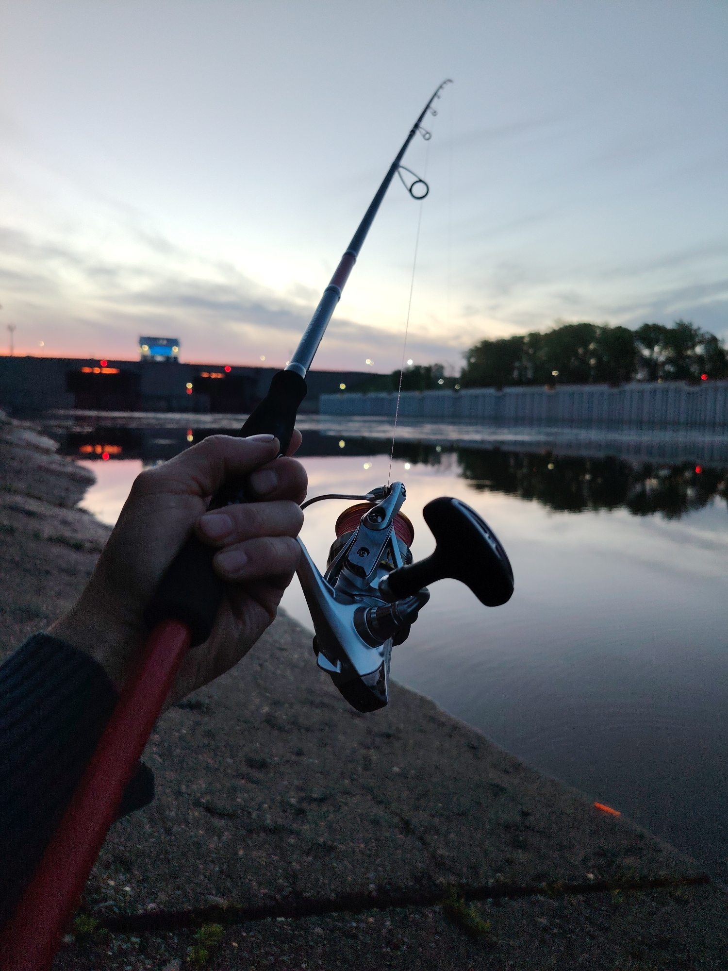 A person holds a fishing rod in their hand, with a silver fishing reel attached, against a backdrop of a body of water at dusk or dawn. The sky is a gradient of blues and light oranges, reflecting in the water. In the background, there are structures that suggest an urban setting, possibly a dam or reinforced embankment with buildings and lights visible. Trees line the upper part of the embankment. The location is likely the Kama Reservoir in the city of Chaikovsky, Russia, based on the presence of the reservoir and the type of structures seen along its banks. The person is standing on a concrete or stone structure, part of the embankment, casting a line into the water.