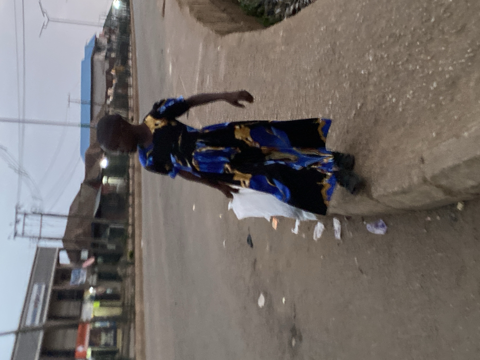 A woman is standing on the shoulder of a road in Bukuru, Nigeria. She is wearing a dress with blue, black, and gold patterns. A white plastic bag and scattered trash surround her feet. Buildings are visible in the background, with electrical lines overhead. The sky is overcast.