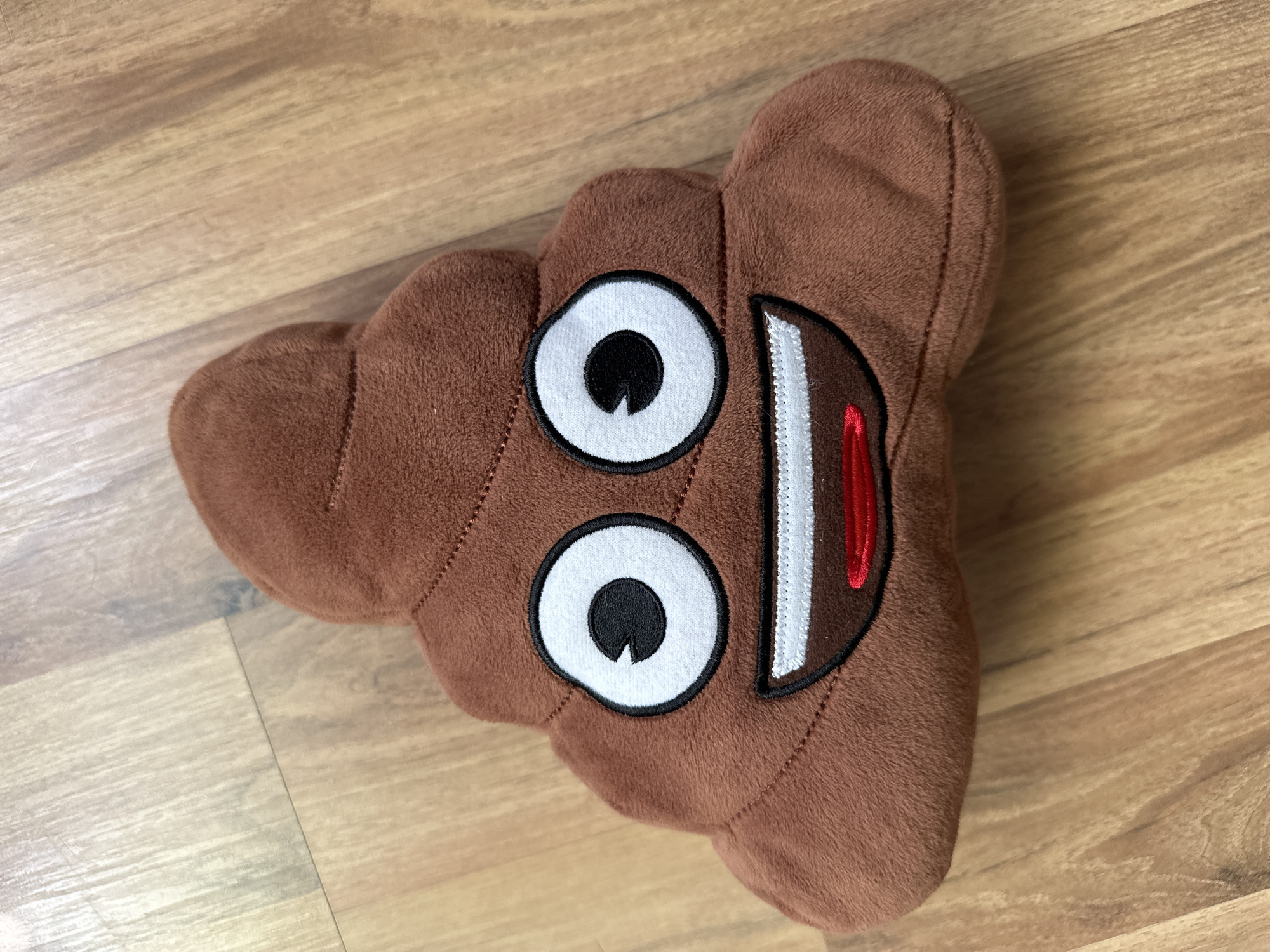 A brown, plush toy shaped like the "poop emoji" rests on a light brown wooden floor. The toy features two large, white circular eyes with black pupils and small white crescent highlights. Below the eyes, a wide, open smile is depicted with white embroidered teeth and a red tongue. The plush material is a uniform shade of brown and has visible stitching delineating the swirled shape of the emoji and outlining the facial features. The wooden floor consists of multiple planks exhibiting variations in grain and color, with distinct seams visible between them. Overhead lighting reflects subtly off the polished surface of the floor.
