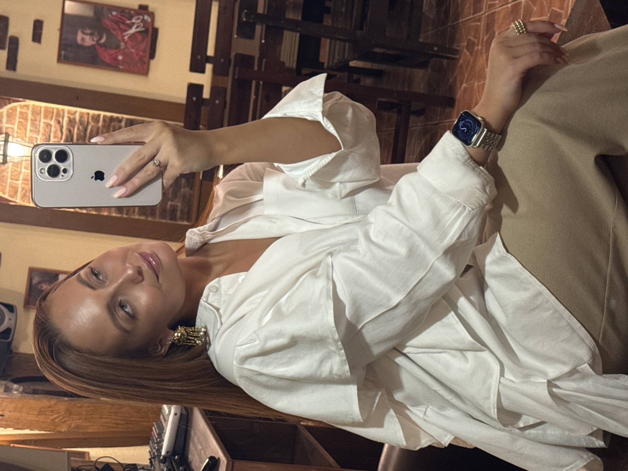 A woman with long, auburn hair is taking a selfie in a dimly lit room. She is wearing a white, oversized collared shirt with layered ruffled detailing on the sleeves and a tan-colored skirt. She has on large gold earrings, a silver smartwatch, and a gold ring. Her left hand is holding up a silver iPhone 14 Pro to take the picture. In the background, there are wooden chairs and tables, a framed picture of a man in a red outfit on the wall, and some audio equipment on a shelf. The overall ambiance suggests a casual setting, possibly a bar or restaurant. The location is identified as Chaikovsky, Russia.