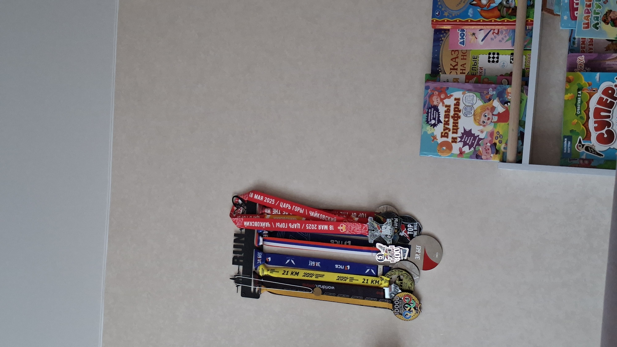 The image shows a collection of running medals hanging on a light-colored wall, next to a shelf with children's books. The medals are stacked vertically, each attached to a ribbon. The top medal has a red ribbon with Cyrillic text stating "18 May 2025 / Tsar of the Mountain / Chaikovsky". Another medal features a black-and-white image of a hill and the words "TOP OF THE HILL". Several medals are associated with 21km races and have ribbons with varying colors, including yellow, blue, and red, white, and blue. The bottom medal has "10000 METERS" written on it.
The bookshelf to the right contains children's books with titles such as "Letters and Numbers", and "Super". It is difficult to determine any interactions or presence of people in the scene from the given information. The environment appears to be inside a home or possibly a school/daycare.