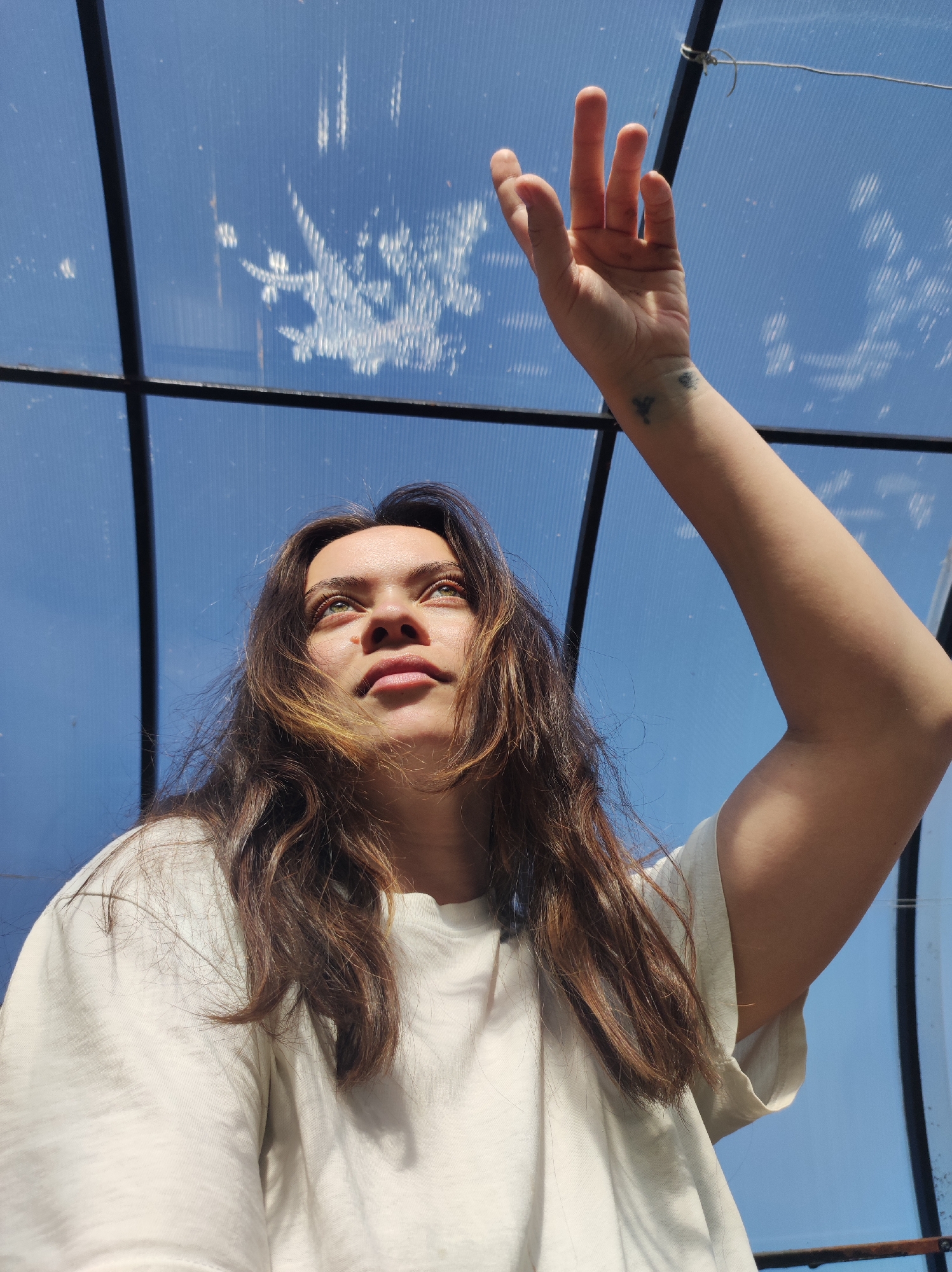 A low-angle shot captures a young woman with light skin, green eyes, and long brown hair, partially highlighted with blonde streaks, looking upwards towards a light blue ceiling. She is wearing a light-colored t-shirt. Her right arm is raised with her hand outstretched, fingers spread, seemingly reaching towards the ceiling. A small tattoo is visible on her right wrist.
The background consists of a grid-like structure with black bars, forming the framework for a translucent, light blue ceiling material. The ceiling has some opaque marks on it, possibly dirt or water stains. The location, Фоки, Russia, is not verifiable from the image.