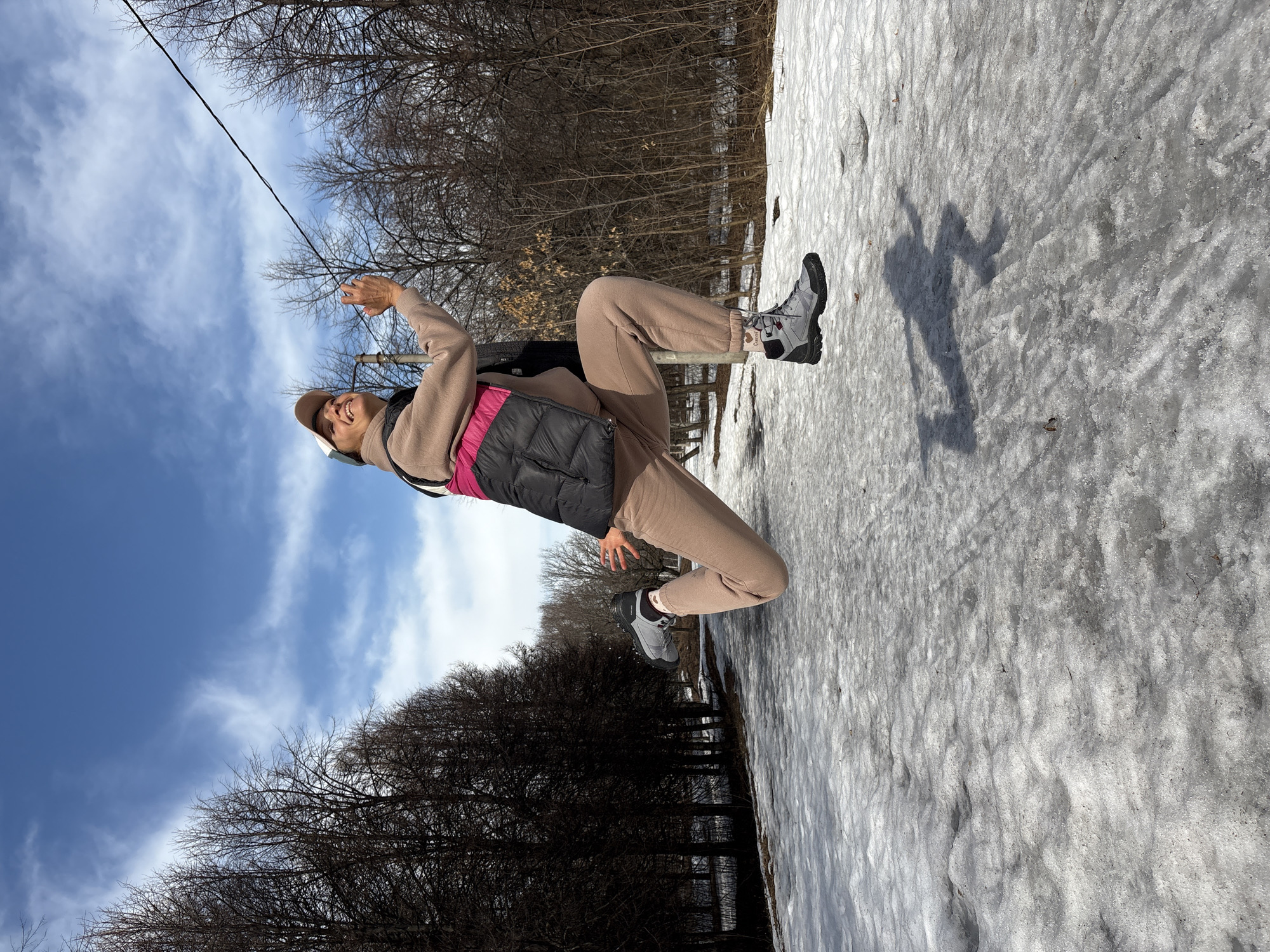 A person wearing a tan outfit, gray boots, a brown hat, and a dark vest with a pink lining is posing outdoors. The person is standing in a snowy environment, with trees and a blue sky visible in the background. The scene appears to be in a park, as evidenced by the trees and the presence of snow. The person is smiling and holding one hand up, as if reaching for a wire in the sky. The ground is covered in slushy snow. The location is Ulyanovsk, Russia.