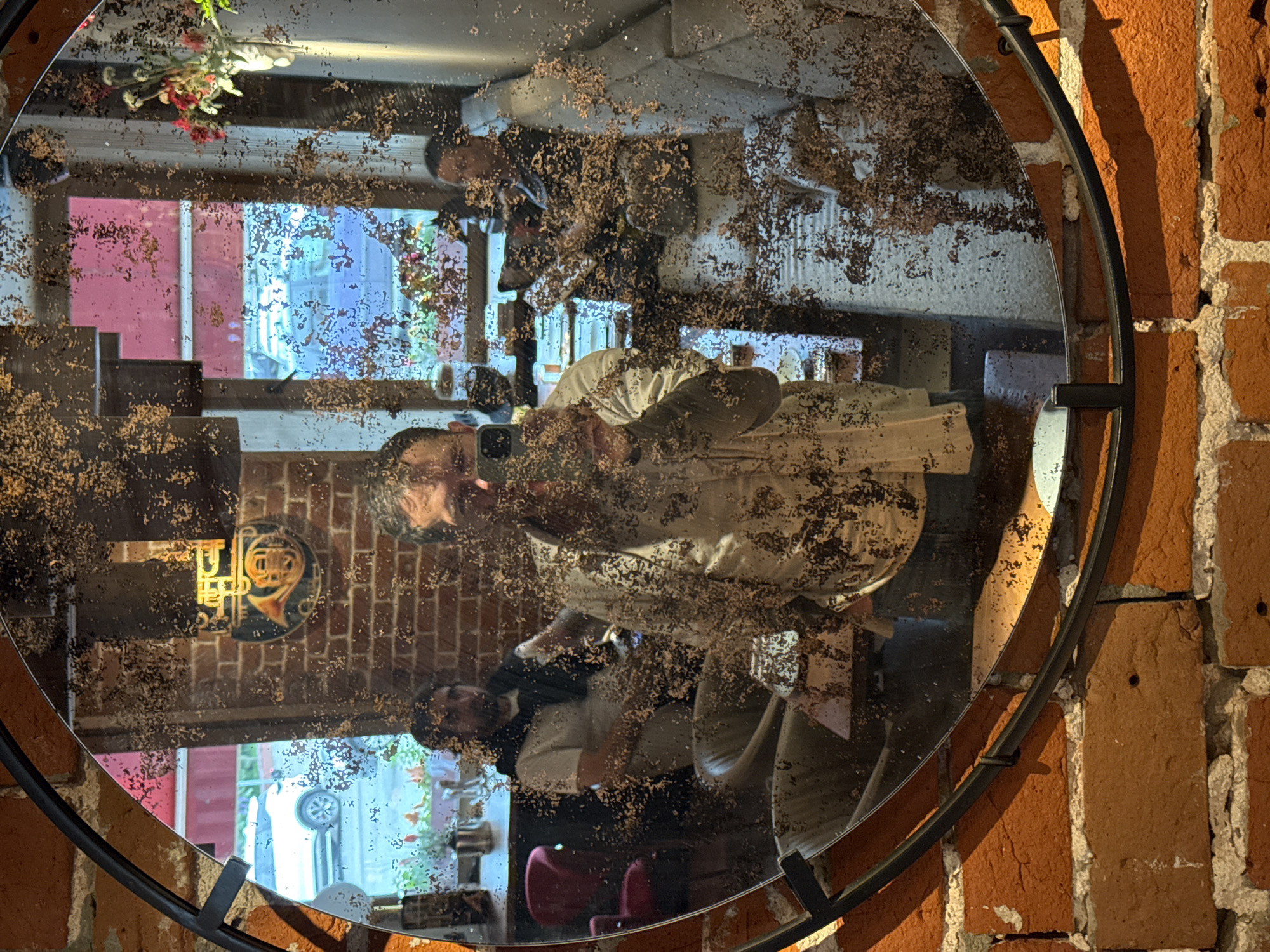 A man is taking a selfie in a round, distressed mirror. He is in the foreground, reflected in the mirror, holding a smartphone up to take the photo. He is wearing a light-colored, possibly cream, shirt with a dark, textured pattern. He has short, dark hair and a beard. The mirror is dirty and appears to have a lot of dirt or debris on its surface, obscuring some of the reflection.
In the background, reflected in the mirror, are two other people. To the left, a person wearing a white t-shirt and a dark scarf draped over their shoulders sits in a chair. To the right, another person wearing a dark shirt is visible, also seated.
The environment appears to be an indoor space, possibly a cafe or restaurant, with brick walls visible both in the foreground and reflected in the mirror. There are windows in the background, showing a street scene with a white car parked outside. A decorative item resembling a French horn is mounted on the brick wall behind the man taking the selfie. Red chairs are also visible in the reflected space. The overall impression is a candid moment captured in a somewhat worn or vintage-styled setting. There is no information to suggest this location is in Nizhny Novgorod, Russia.