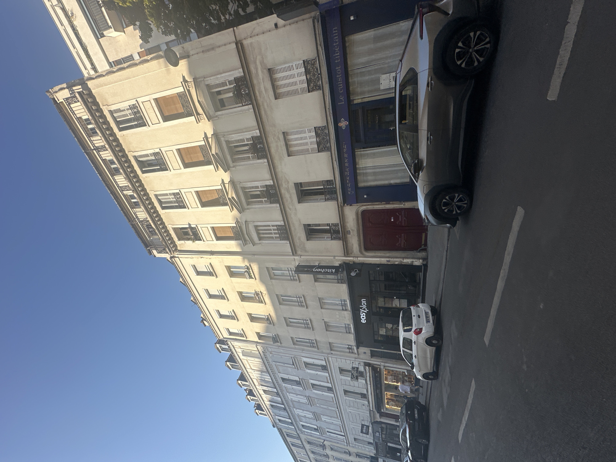 A low-angle, full outdoor shot shows a Parisian street with buildings on the left and right of the frame. The buildings are old and have a light yellow facade. On the left, there is a building with many windows and balconies. A sign for "easy plan" is visible on the ground floor. A white car is parked on the street in front of the building. On the right, a darker facade building has a blue awning with text in what appears to be Tibetan script and "Le cuistot tibetan" written in white. A silver crossover car is parked in front of this building. The sky is clear and blue.