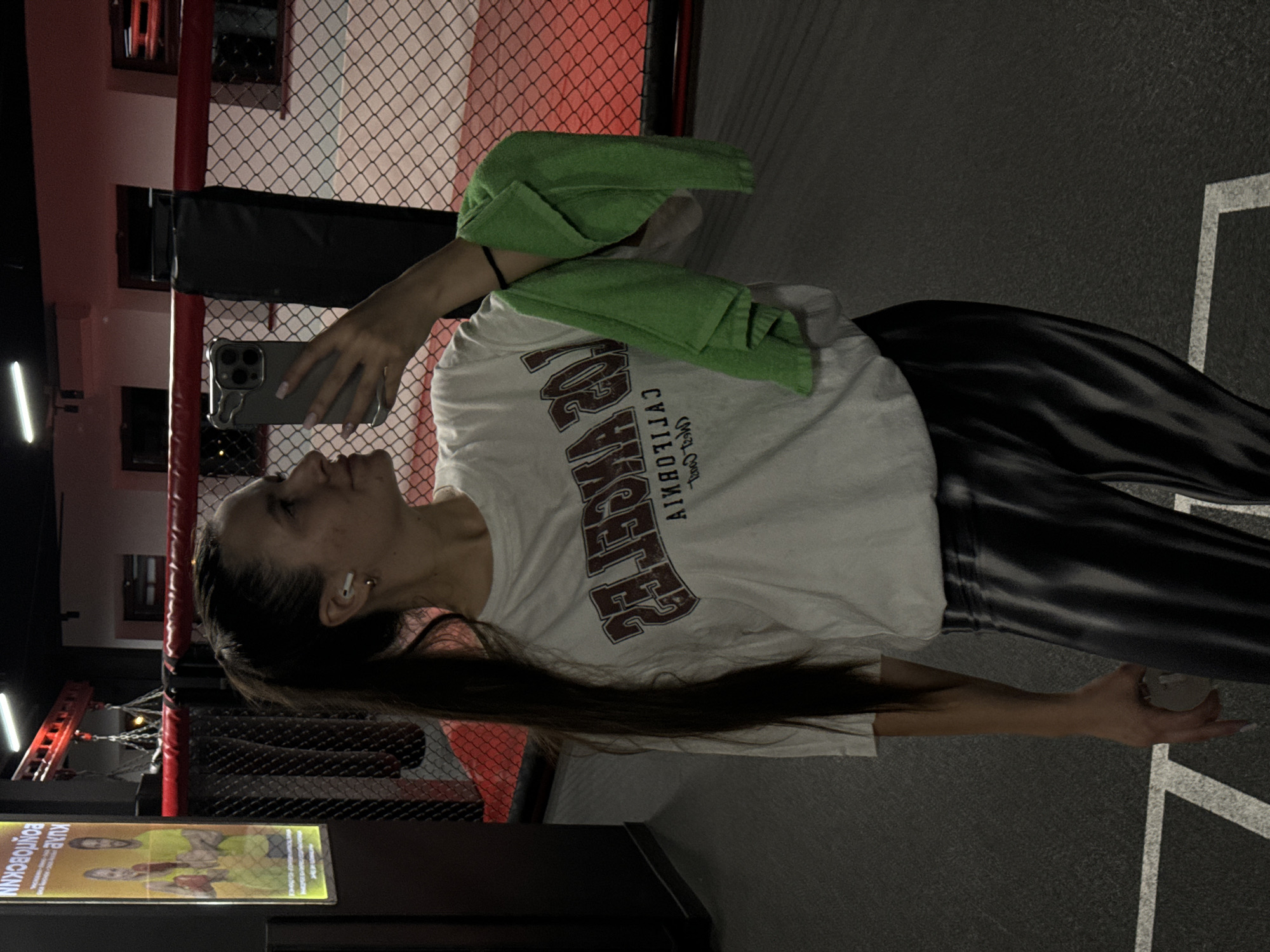 The image depicts a woman taking a selfie indoors in what appears to be a boxing gym or mixed martial arts facility, presumably located in Vniissok, Russia. The woman is wearing a white t-shirt, black and white striped leggings, and has a green towel draped over her shoulder. She is holding a smartphone up to take a selfie. In the background, there is a boxing ring with red corner posts and a chain-link fence. There are other gym-related items visible in the frame.